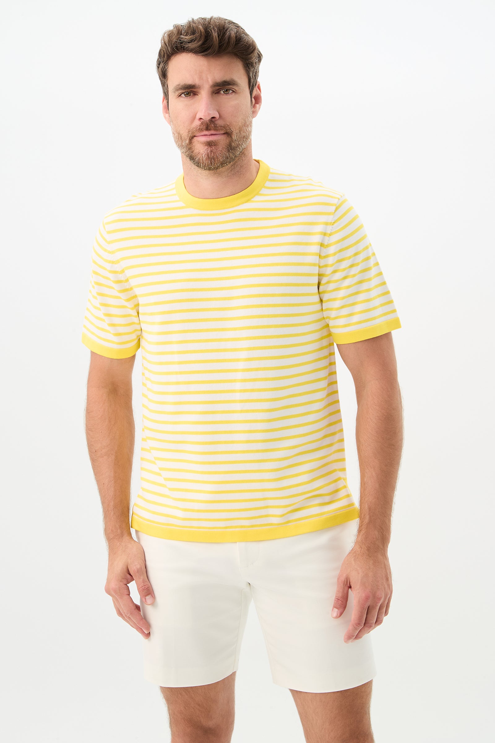 A man with short brown hair and a beard wears the Giovanni Stripe Short-Sleeve Pullover in Desert Sun/Whitewash and white shorts, standing against a plain white background.