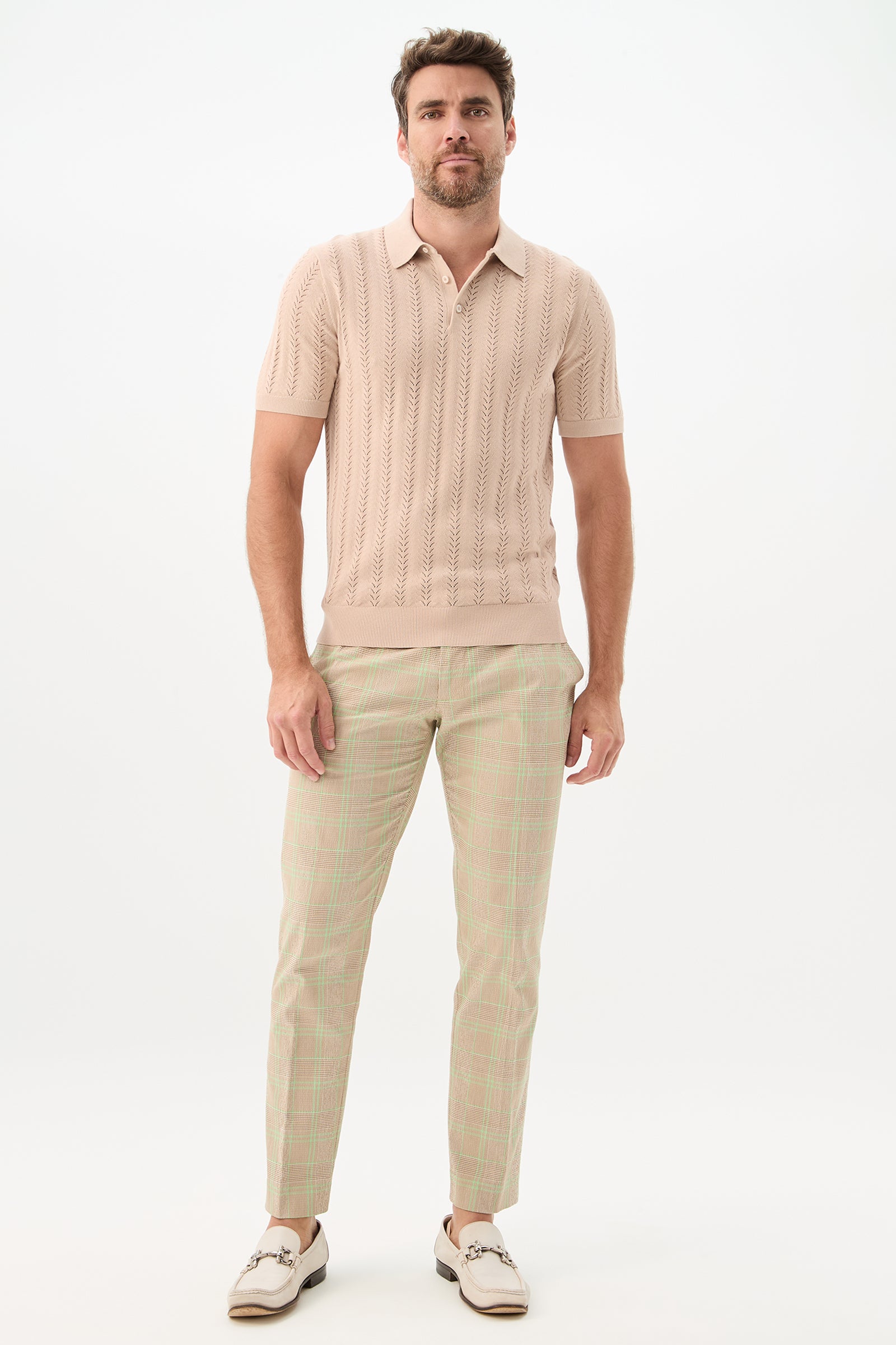 A man with short brown hair and a trimmed beard stands against a white background wearing a beige short-sleeve knit polo, the Clyde Slim Trouser - Zianieri Plaid, and white loafers with brown accents.