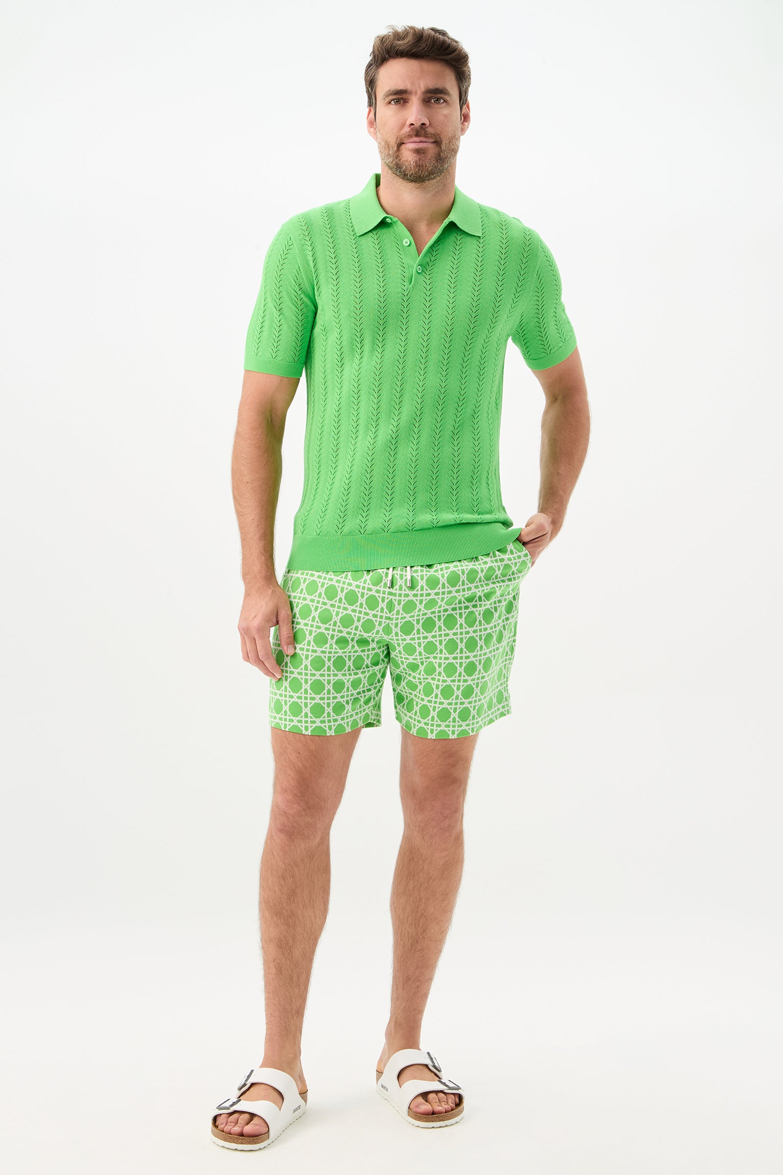 A man with short hair and light facial hair stands relaxed against a white background, wearing bright green shorts (Lounge Short - Billie Jean), a green polo shirt, and white sandals.