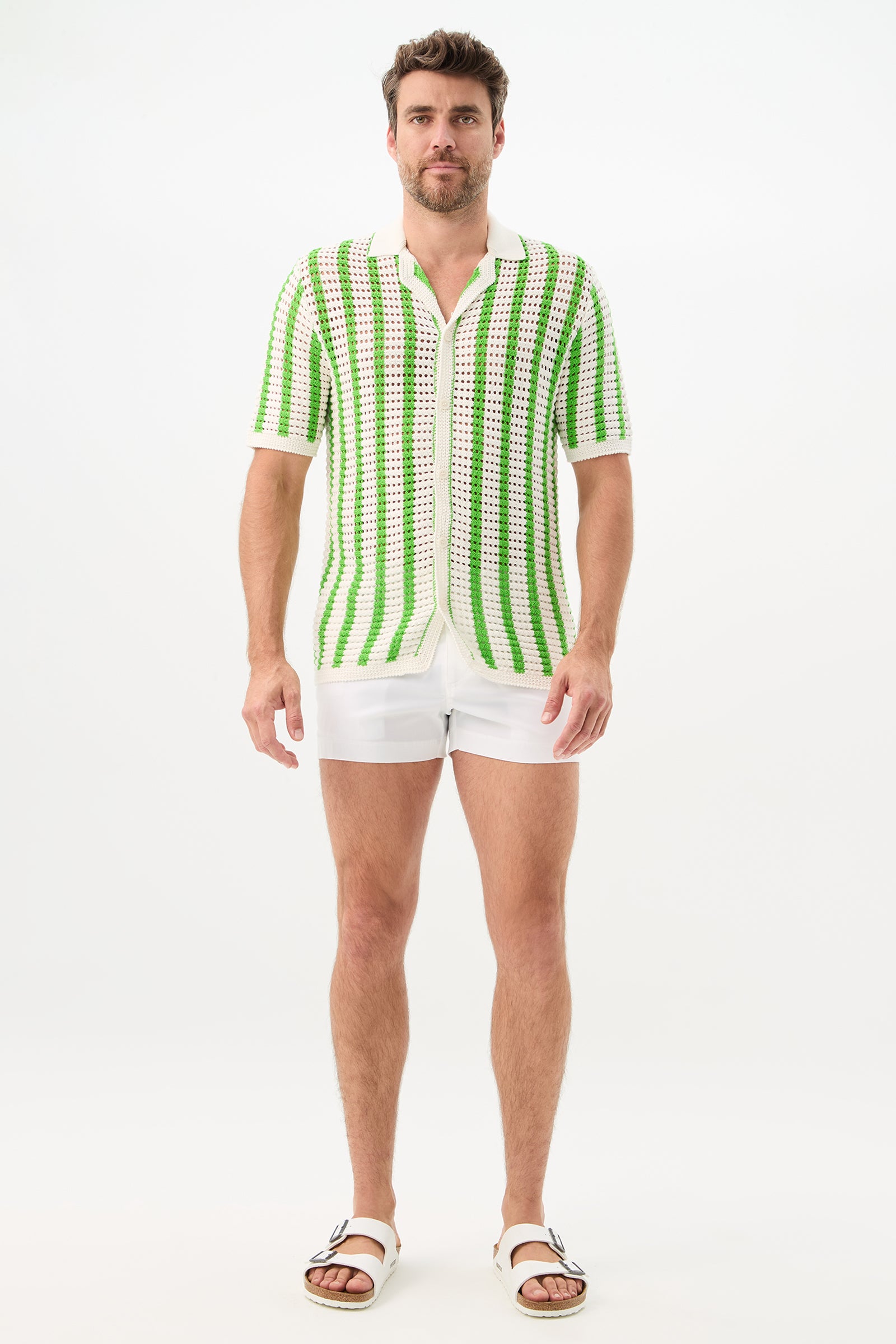 A man with short brown hair and a trimmed beard stands against a plain white background wearing the Easton 2 Short-Sleeve Shirt, which has white and green stripes. He pairs it with white shorts and white sandals.