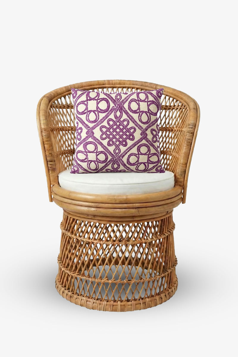 A round rattan chair with a beige seat cushion is accented by the Embroidered Square Pillow, featuring a purple geometric pattern, all set against a plain white background.