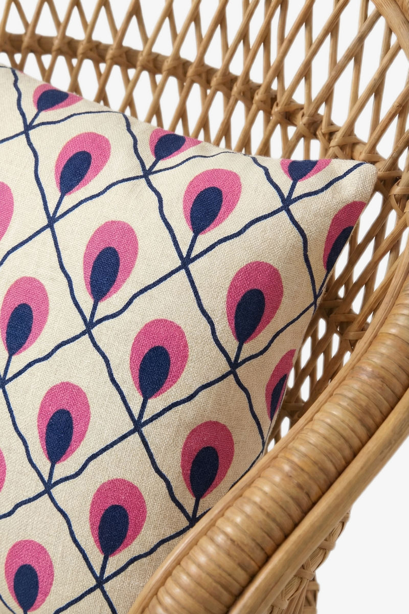 A close-up of a rattan chair with an Embroidered Lumbar Pillow, featuring a geometric pattern of pink and navy teardrop shapes on a cream background.