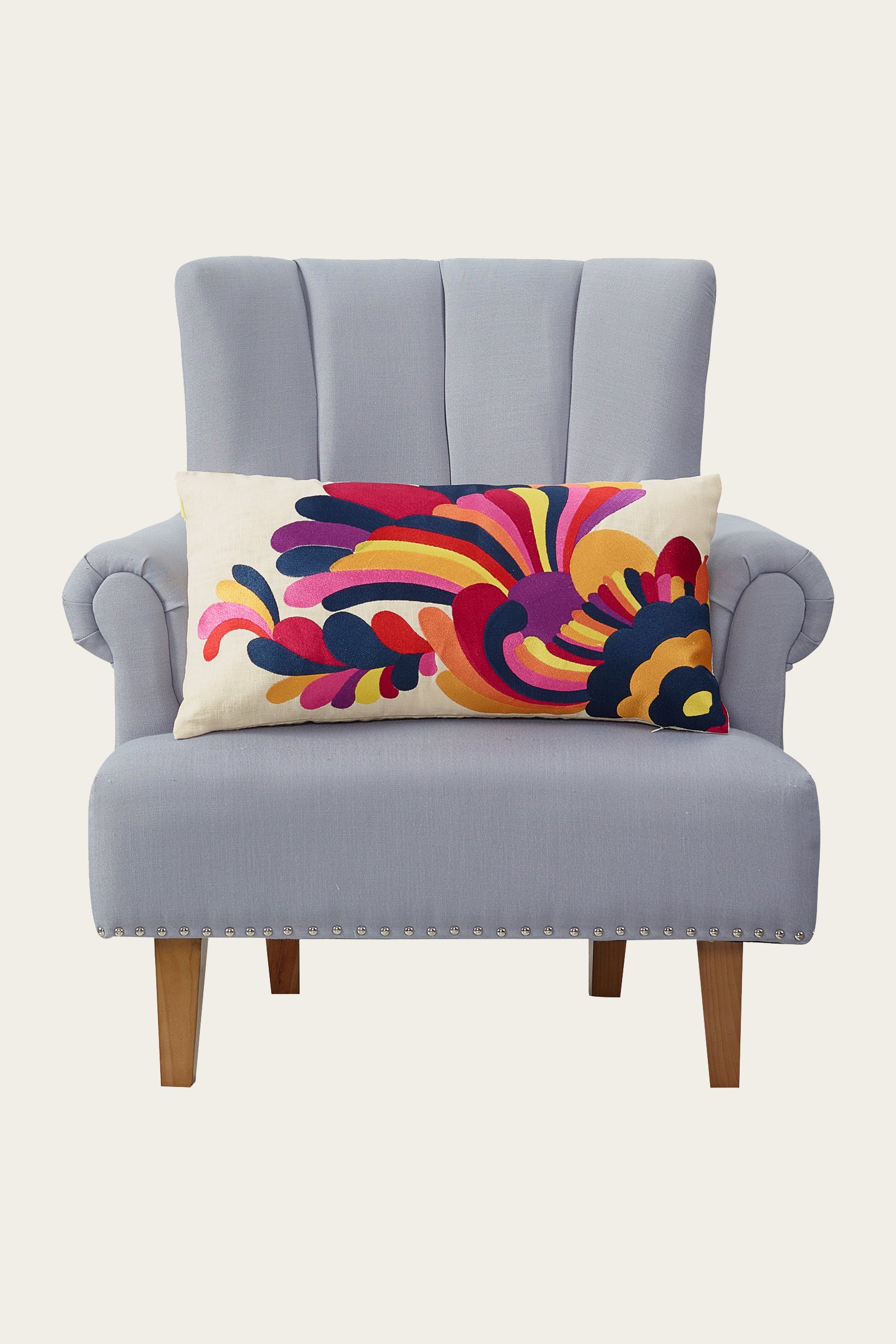 A light gray upholstered armchair with wooden legs, featuring the Escondido Embroidered Throw Pillow in a colorful, abstract pattern of red, yellow, blue, purple, and pink.