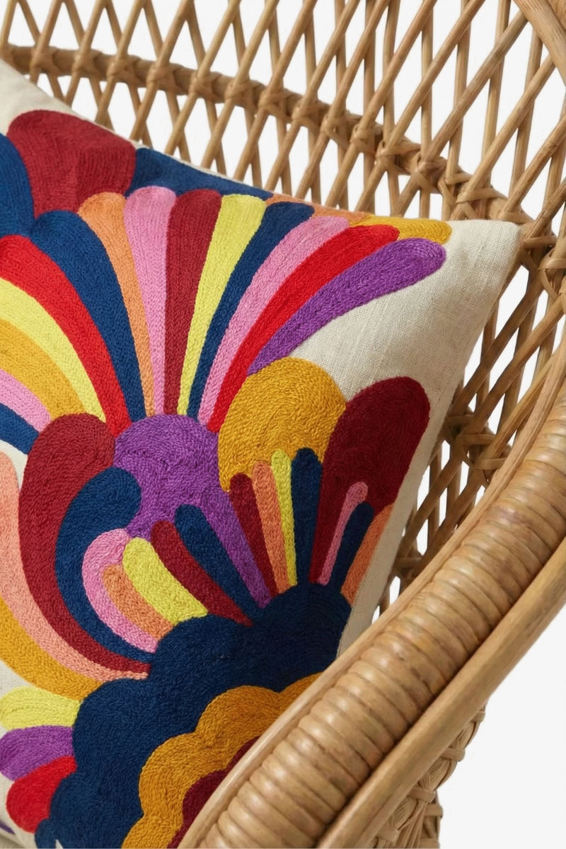 An Embroidered Lumbar Pillow, featuring a white base adorned with colorful, abstract floral patterns in red, yellow, blue, pink, purple, and orange, rests on a rattan chair.