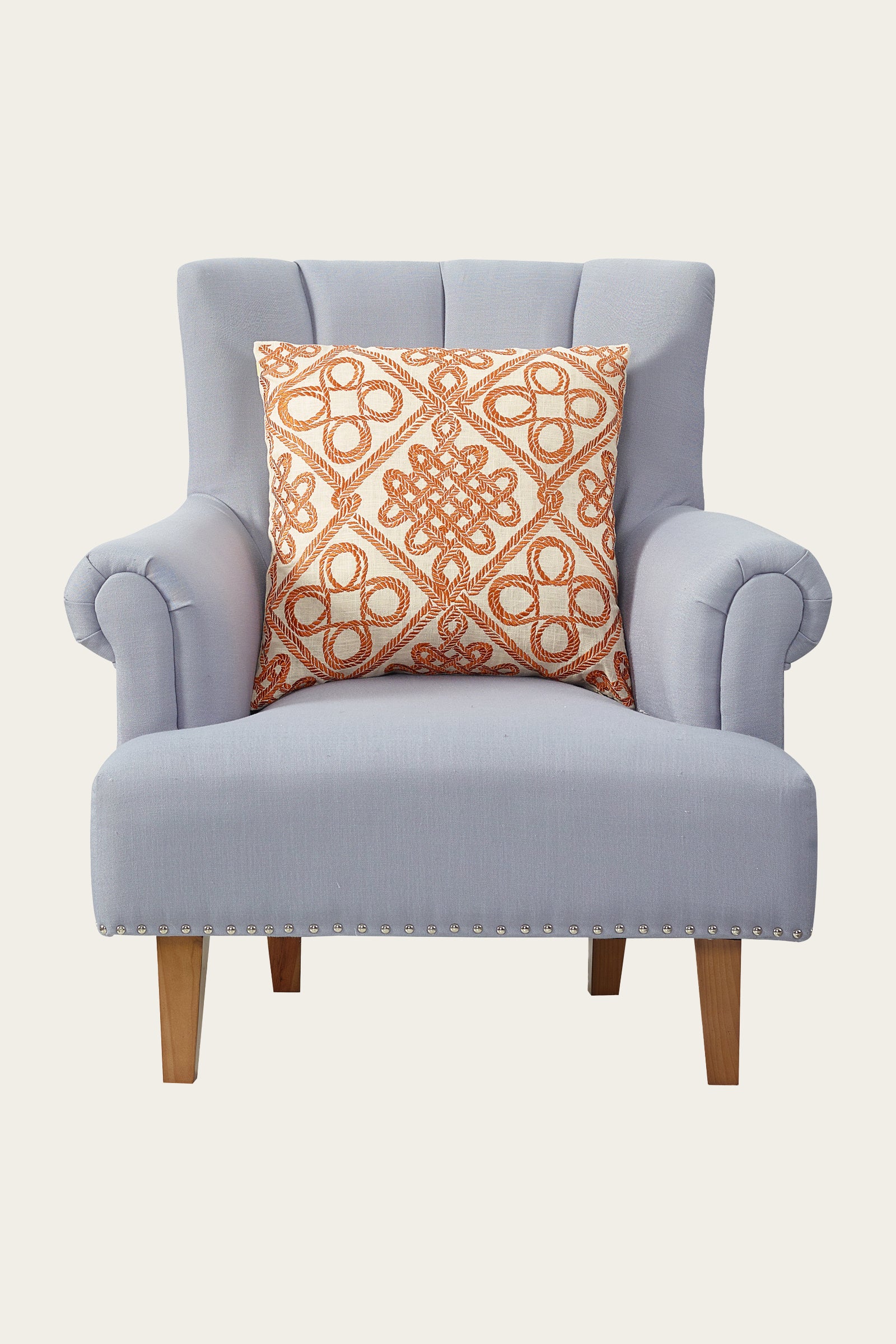 A light gray upholstered armchair with wooden legs and silver nailhead trim features the Costa Mesa Embroidered Euro Throw Pillow in Terra, showcasing an intricate orange and white geometric design.