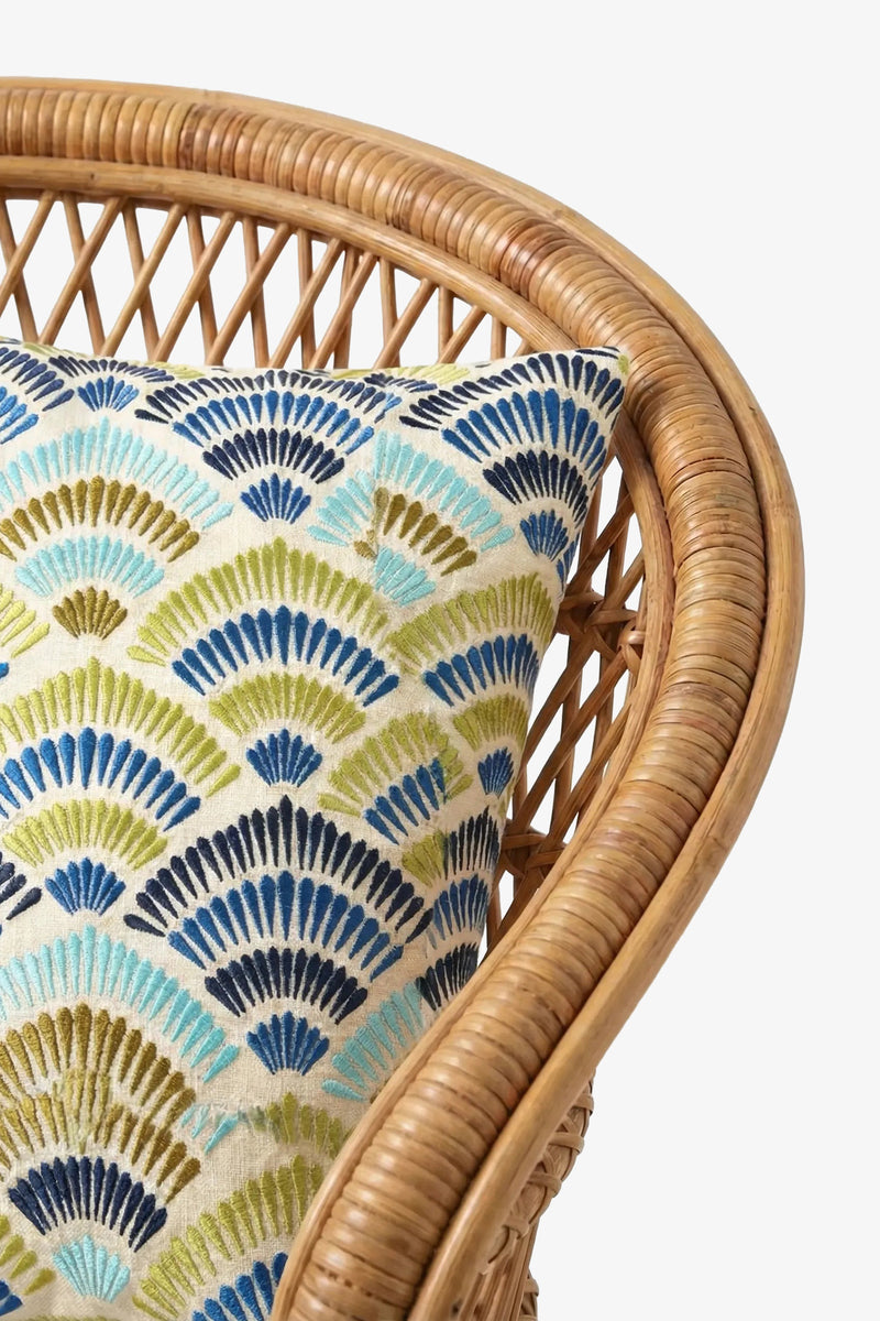 A close-up of a wicker chair with the Pasadena Embroidered Square Pillow, which features blue, green, and teal fan-shaped patterns on a white background.