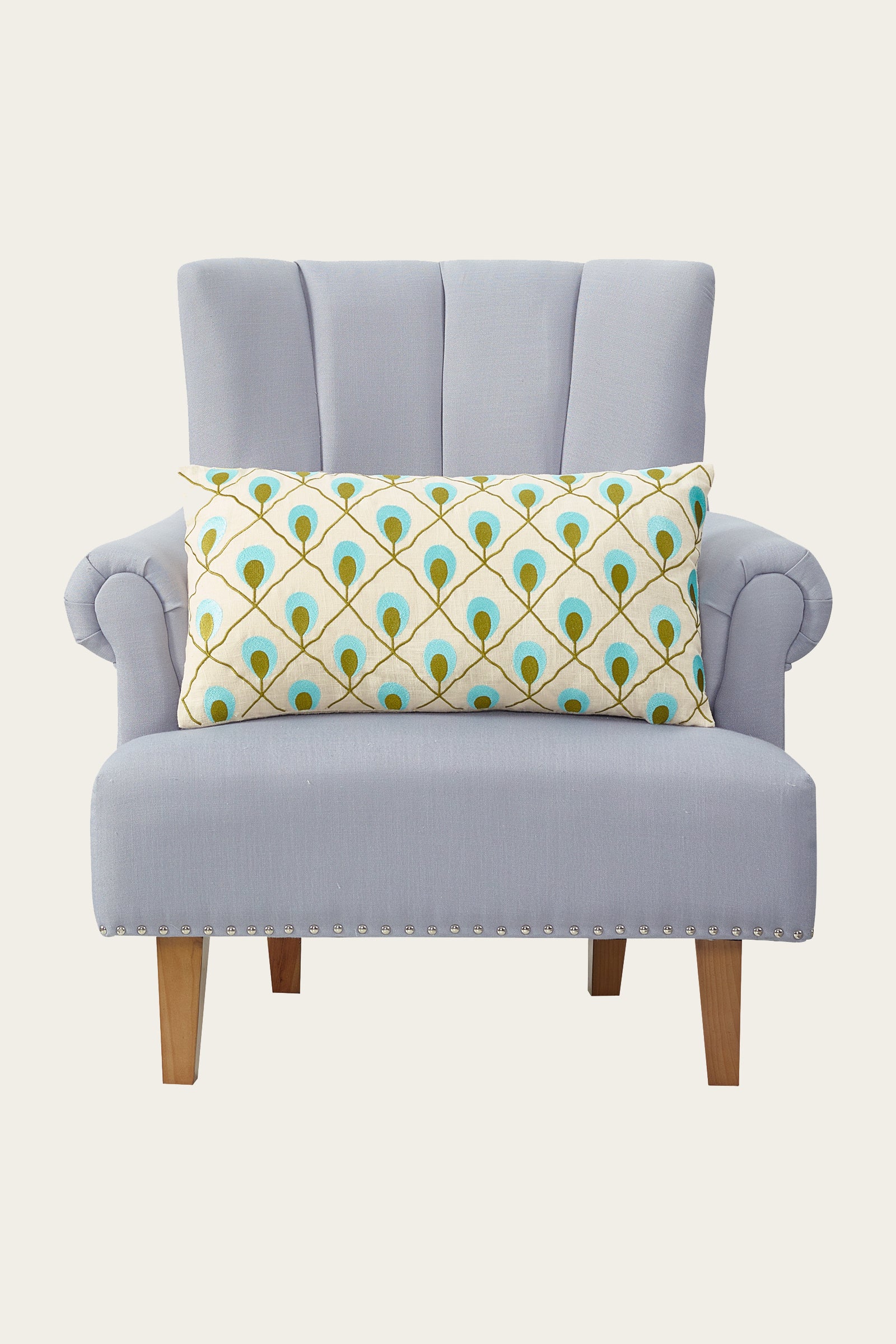 A light gray upholstered armchair with wooden legs, featuring the Roseville Embroidered Throw Pillow in blue, adorned with a cream cover and a peacock feather design in blue, green, and gold.