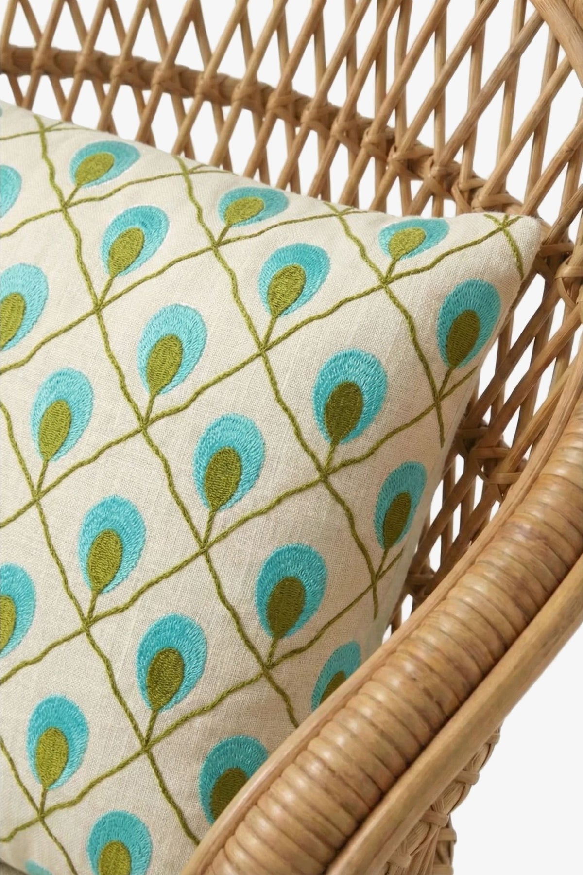 A close-up of a woven rattan chair with a cream cushion, accented by the Roseville Embroidered Lumbar Pillow featuring a blue and green leaf or feather pattern.