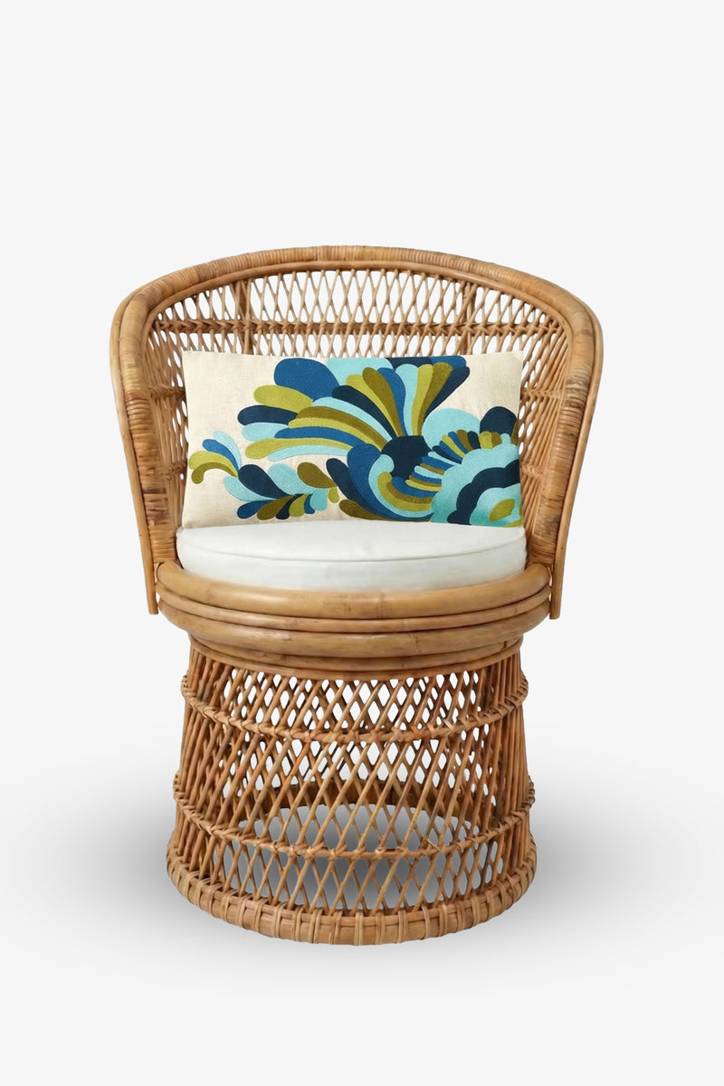 A round wicker chair with an open-weave design and a white seat cushion is paired with the Escondido Embroidered Lumbar Pillow, which features a bold, abstract pattern in blue, green, and yellow tones. The background is plain white.