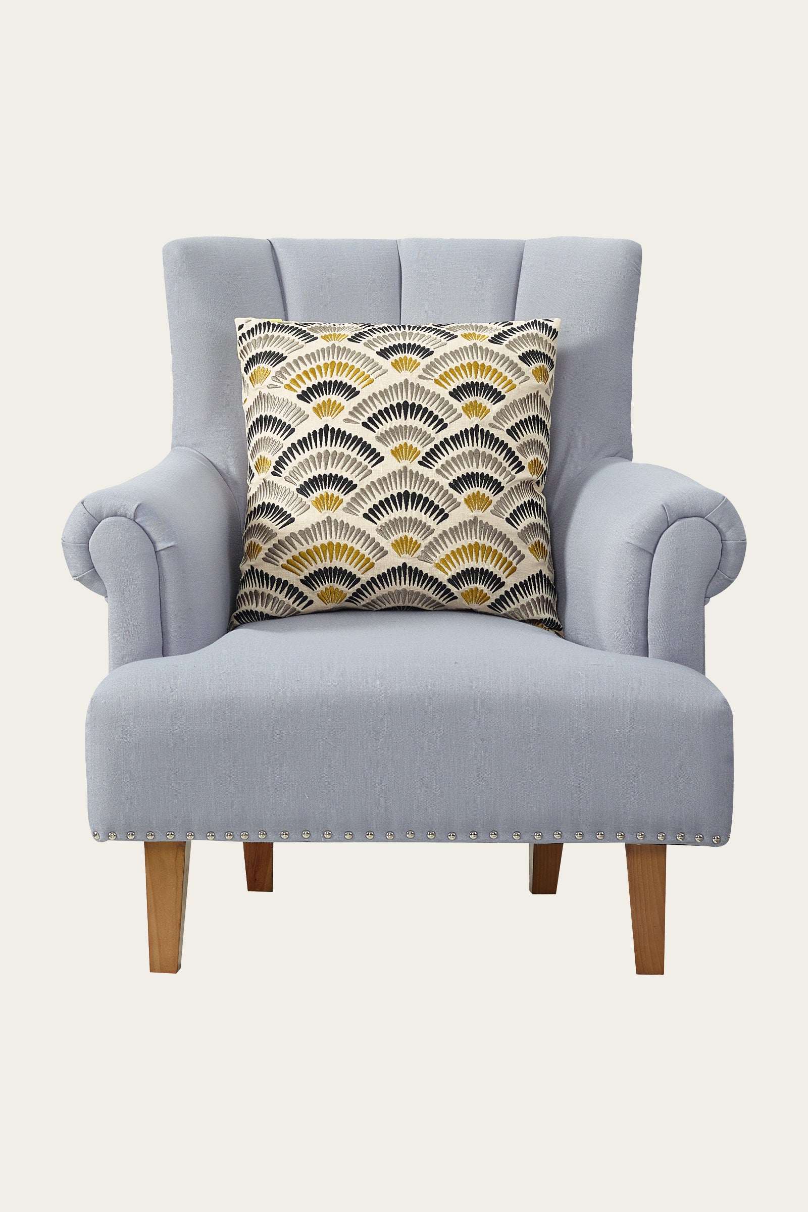 A light gray upholstered armchair with wooden legs features the Pasadena Embroidered Euro Throw Pillow in gold, showcasing a geometric fan pattern in black, white, and gold. The background is plain and cream-colored.