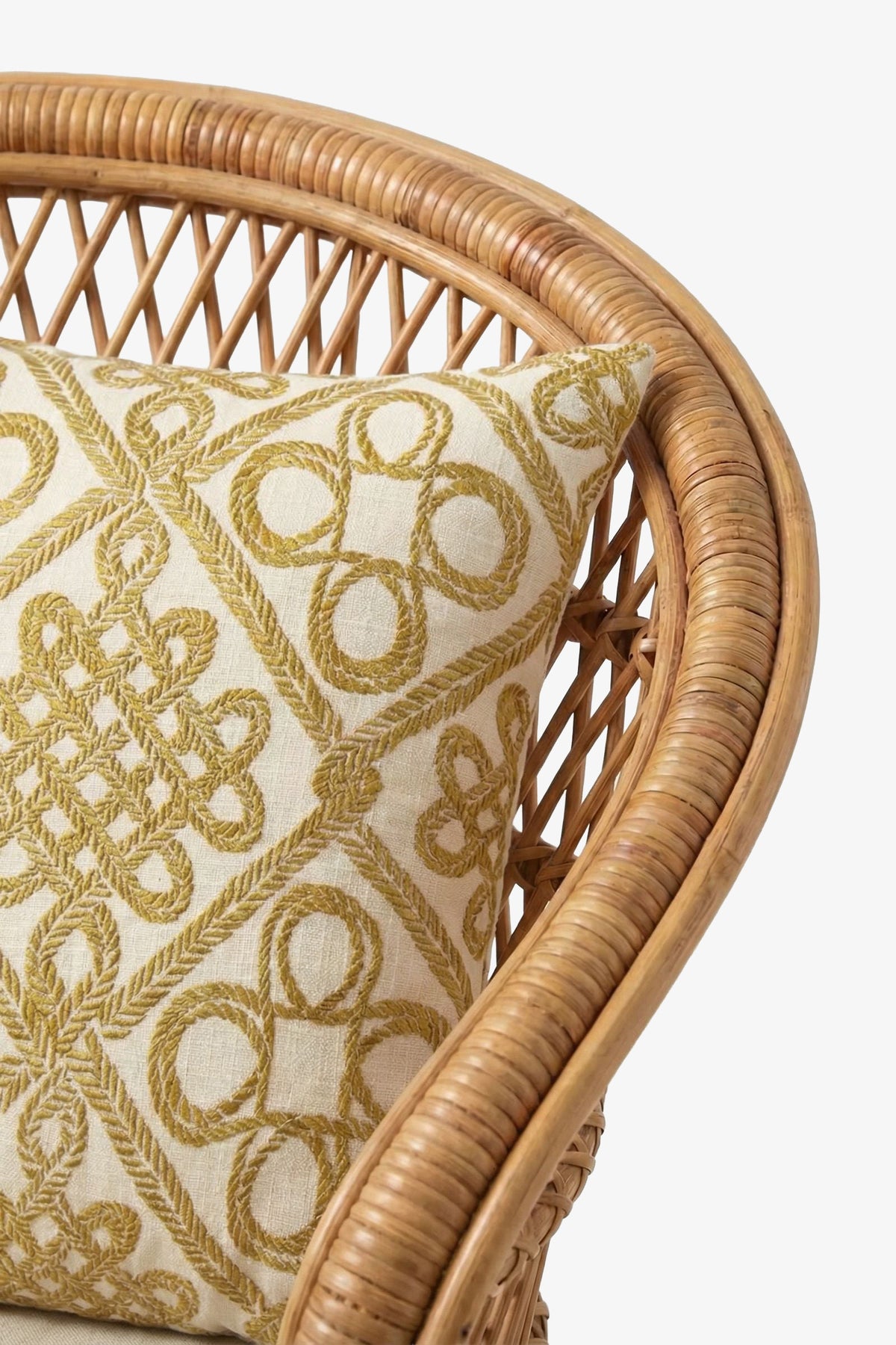 A close-up of a rattan chair with curved armrest, featuring the Costa Mesa Embroidered Square Pillow adorned with gold embroidered geometric patterns on a white background.
