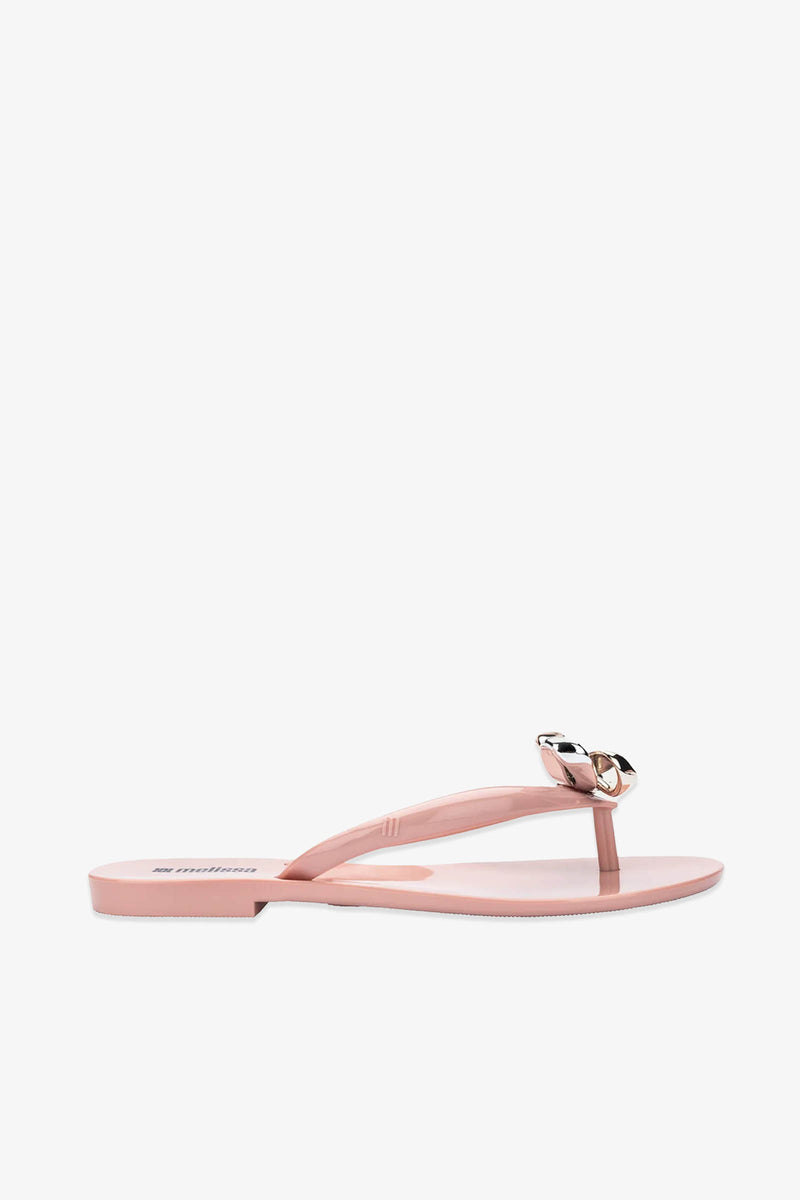 The Melissa Harmonic Eclipse Bow Sandal in pink features a flat thong design with a striking metallic chain detail on the strap, showcased against a white background.