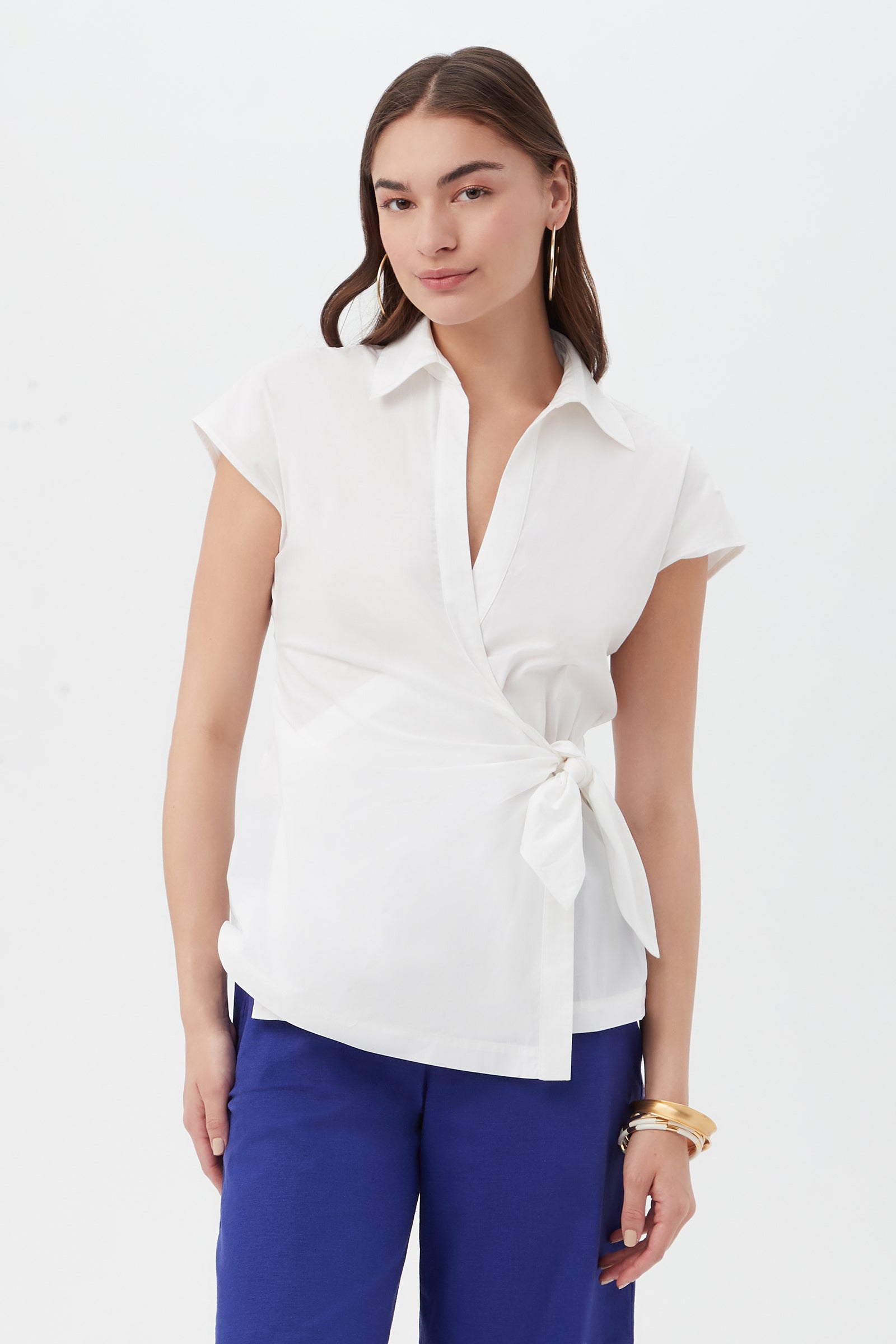 A woman with long brown hair, gold hoop earrings, and bracelets wears the Avant Top in white— a short-sleeve wrap blouse with a side tie—paired with blue pants, standing against a plain white background.