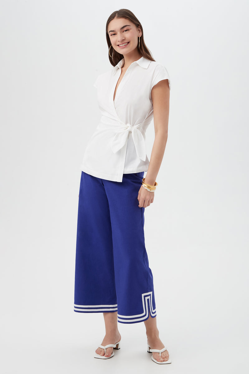 A woman stands smiling in the Avant Top, a white short-sleeve wrap blouse, paired with cobalt blue wide-leg culottes with white-trimmed hems, white sandals, and gold bracelets against a plain white background.