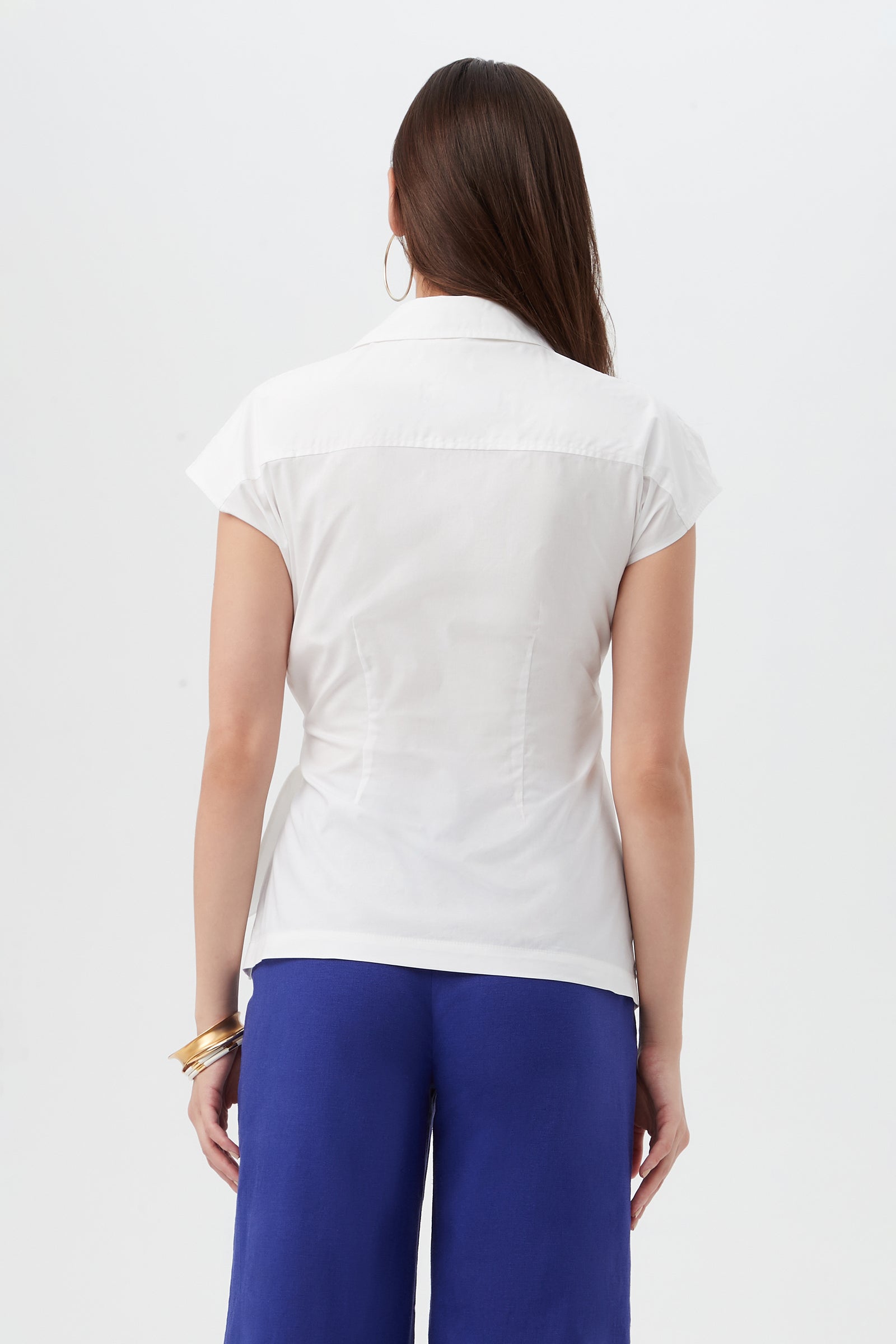 A woman with long brown hair, wearing the Avant Top—a short-sleeved white blouse—paired with blue pants, stands facing away from the camera against a plain white background. She accessorizes with gold hoop earrings and gold bangles.