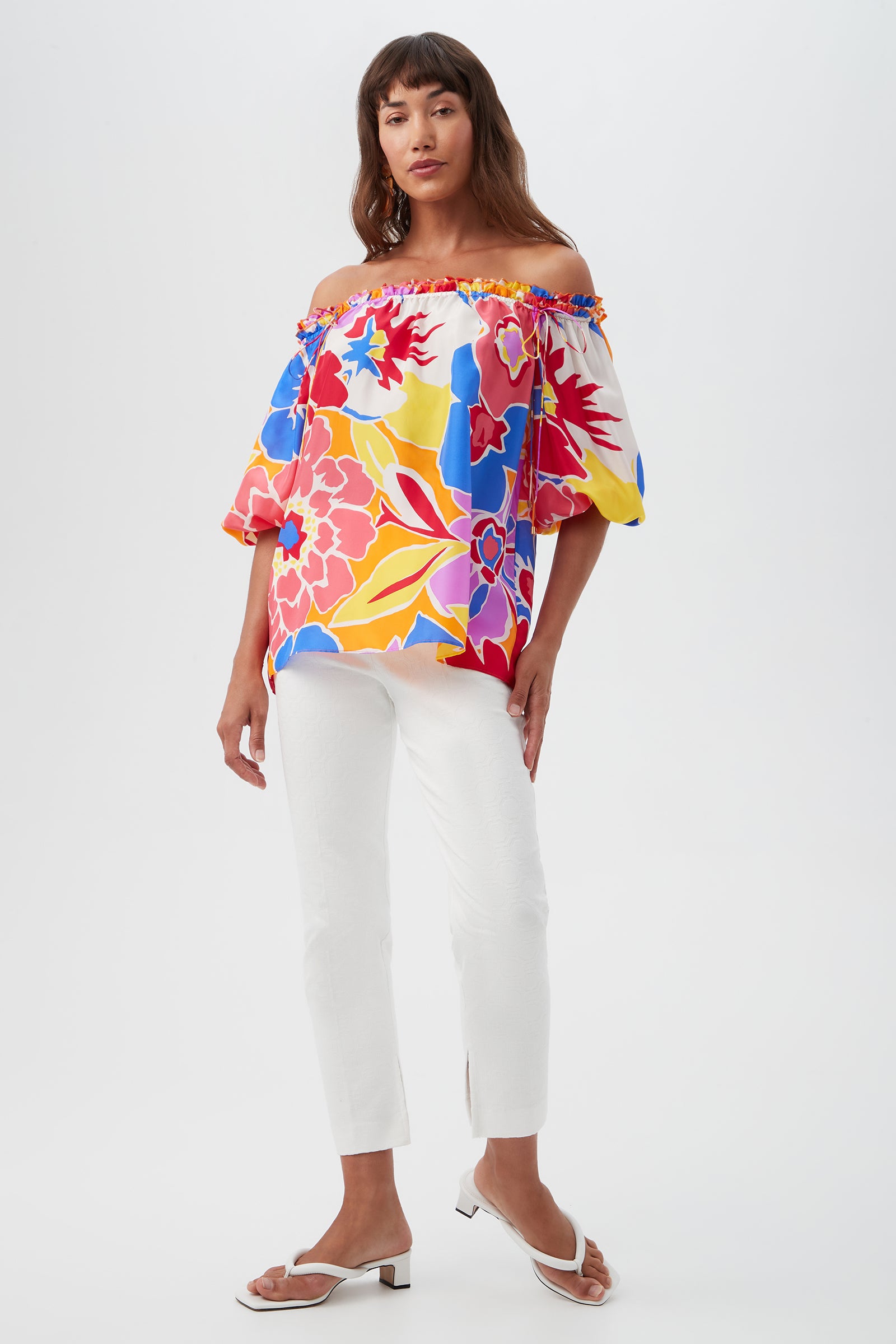 Woman posing in the Jambies Top - Soie Paradis, a colorful off-the-shoulder floral top, paired with white slim-fit pants and white open-toe sandals against a plain light background.