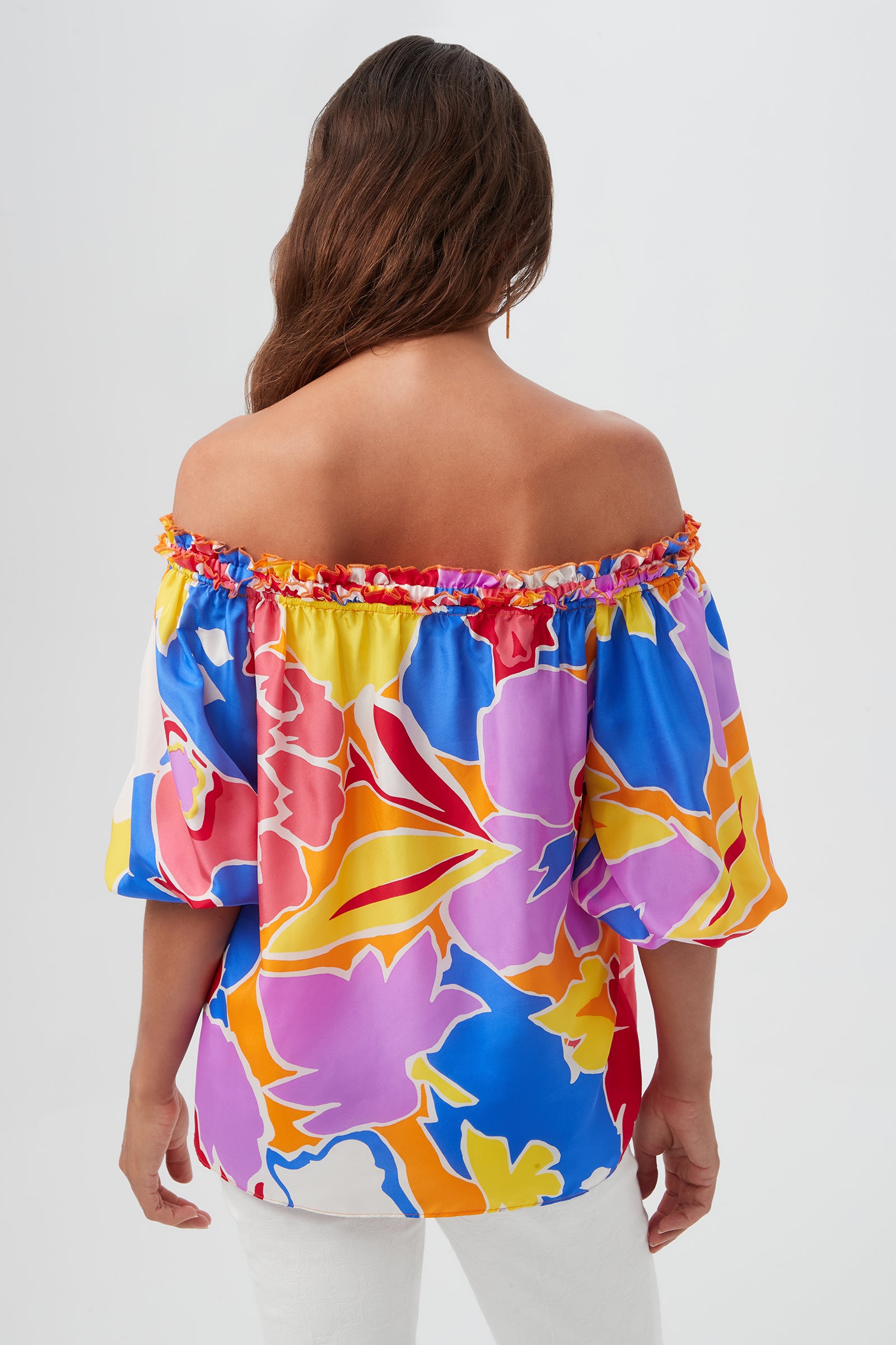 A woman with long brown hair wears the Jambies Top - Soie Paradis, an off-the-shoulder, colorful floral top with puffed sleeves and white pants, shown from the back against a plain light background.