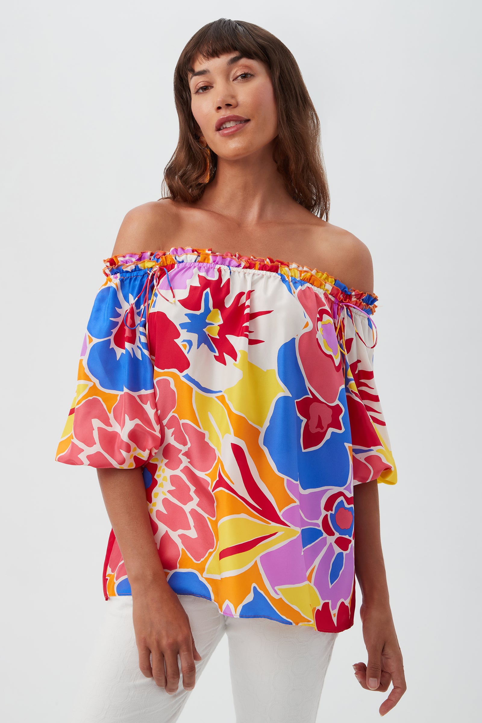 A woman wears the Jambies Top - Soie Paradis, an off-the-shoulder, short-sleeve blouse in a vibrant Soie Paradis floral print, paired with white pants and standing against a plain light background.