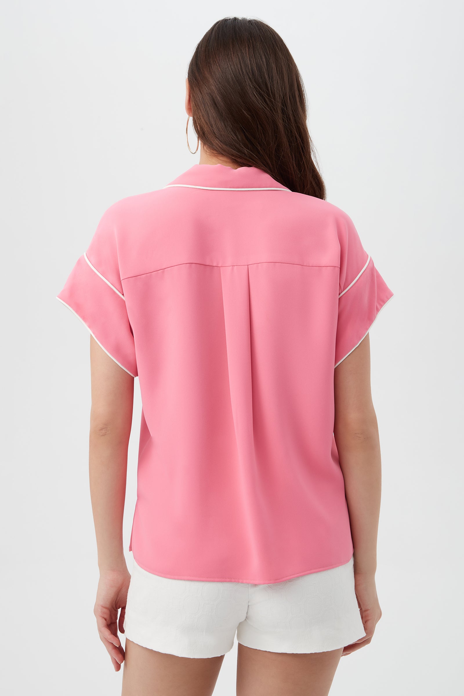 A woman with long brown hair wears the Gideon 2 Top, a pink short-sleeve blouse with white trim, and white shorts, standing with her back to a plain white background.