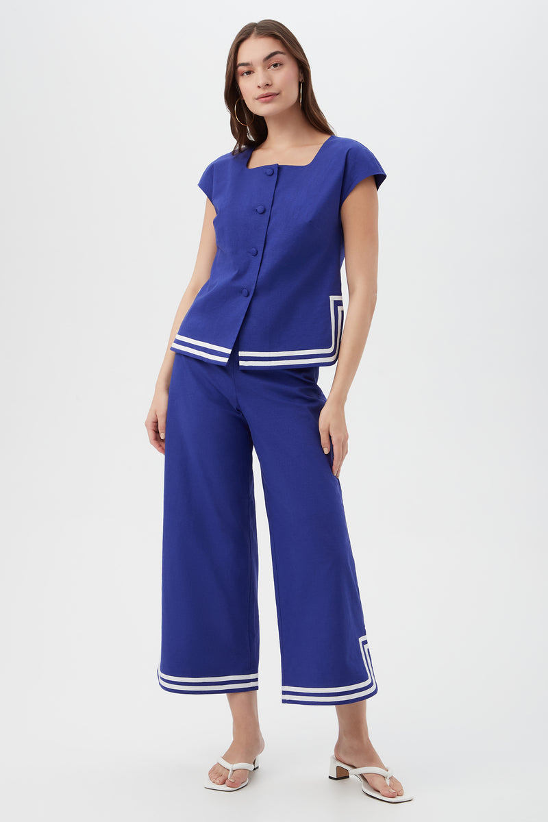 A woman wears the Valence Top, a blue short-sleeve shirt with large buttons and white stripe details near the hem, paired with matching wide-leg pants and white sandals, standing against a plain background.