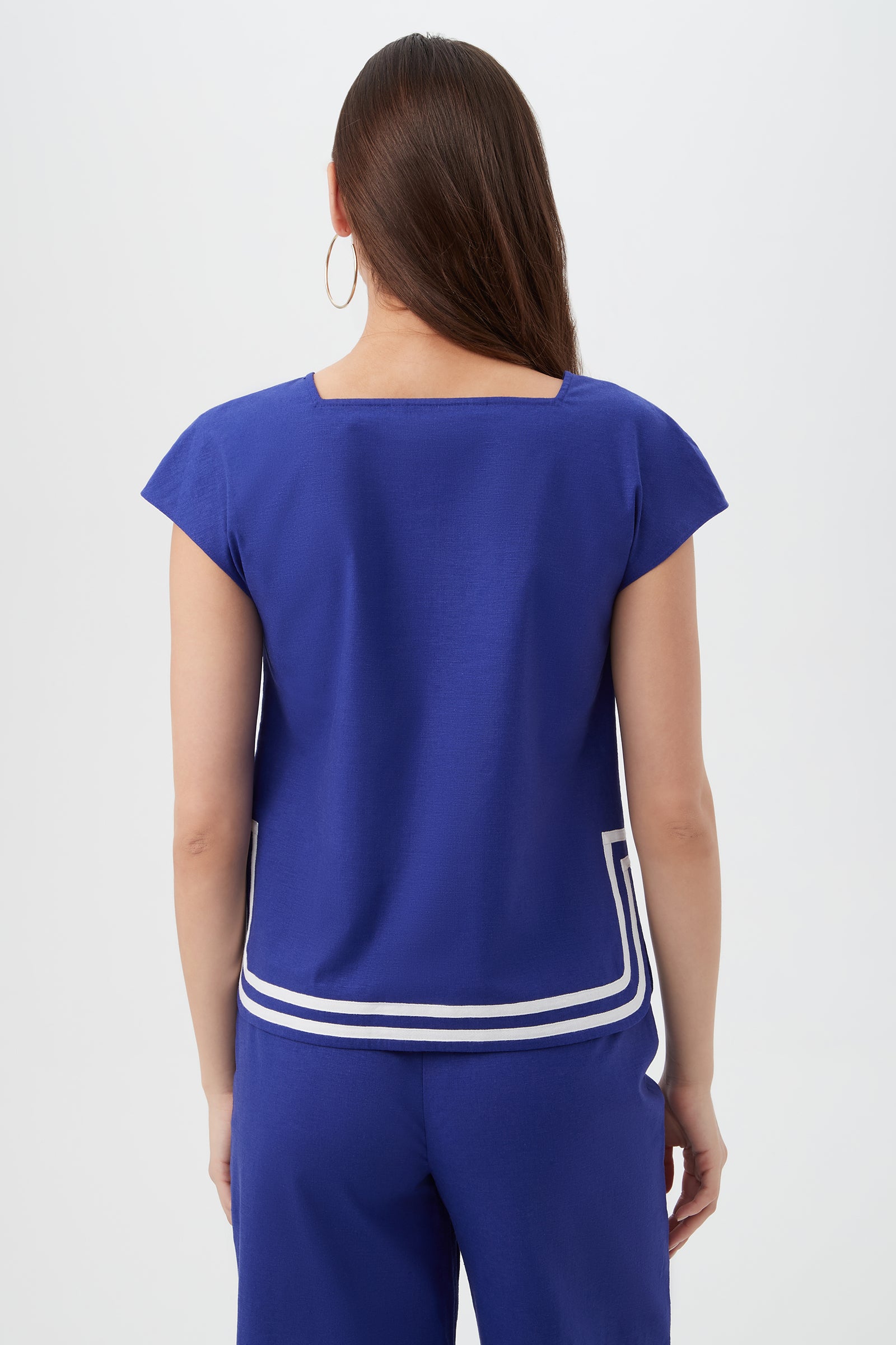 A woman with long brown hair and hoop earrings faces away, wearing the royal blue Valence Top with white trim and matching pants. The background is plain white.