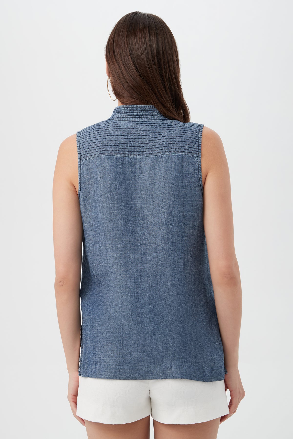 A woman with long brown hair is shown from the back against a plain light background, wearing the sleeveless Lacheri Chambray Top and white shorts.