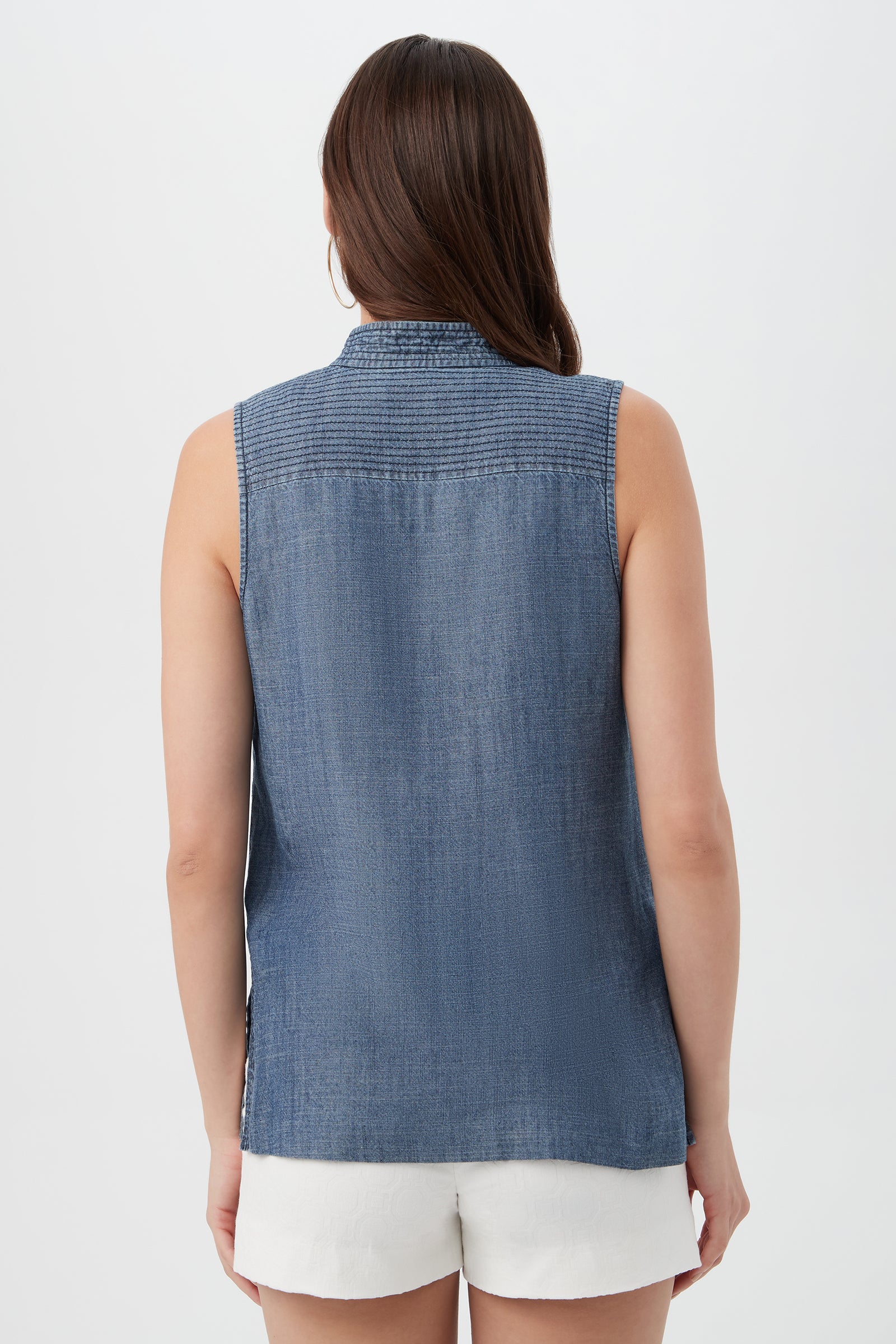 A woman with long brown hair is shown from the back against a plain light background, wearing the sleeveless Lacheri Chambray Top and white shorts.