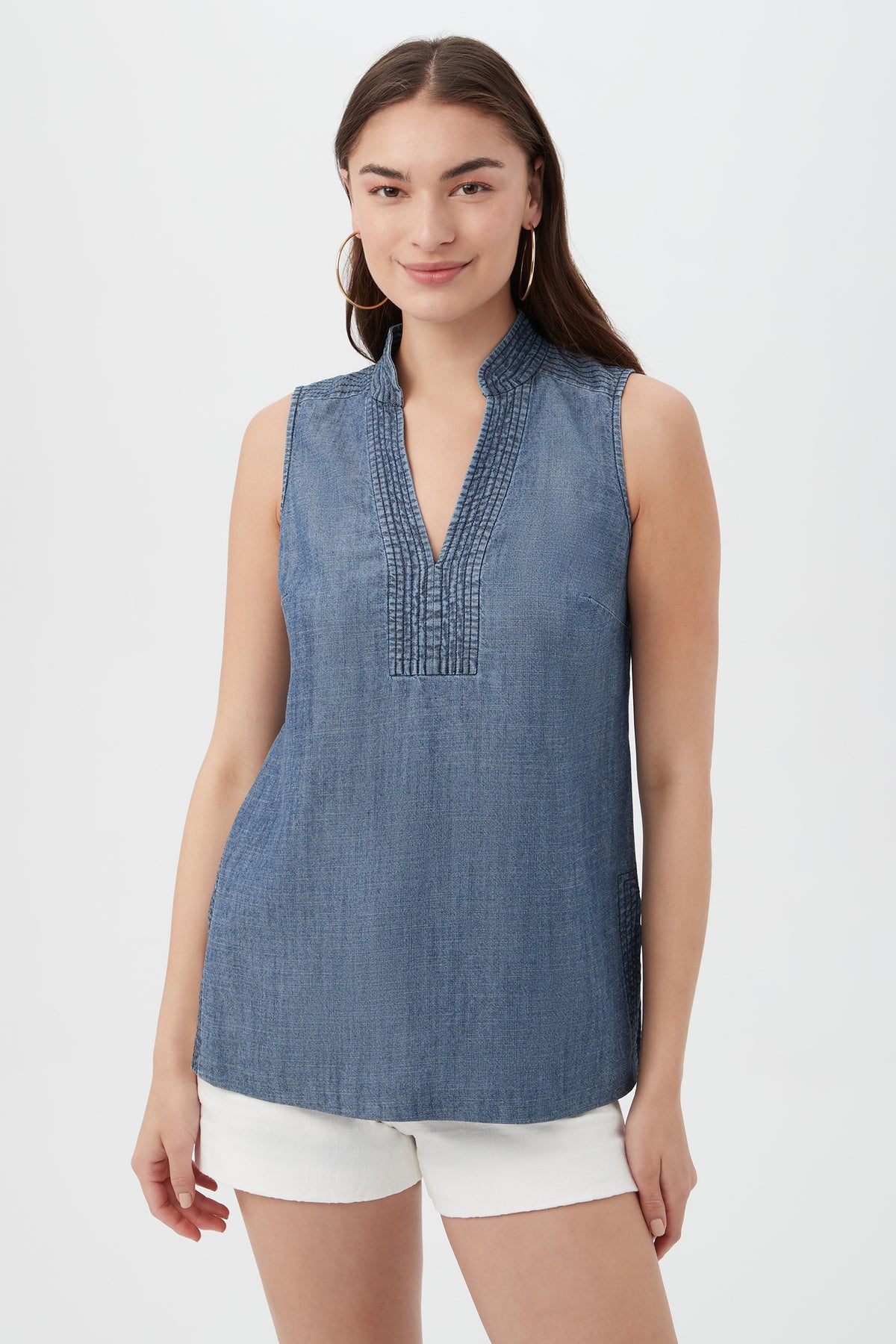 A woman stands against a plain white background, smiling gently at the camera in an indigo Lacheri Chambray Top with a split neckline and sleeveless design, paired with white shorts.