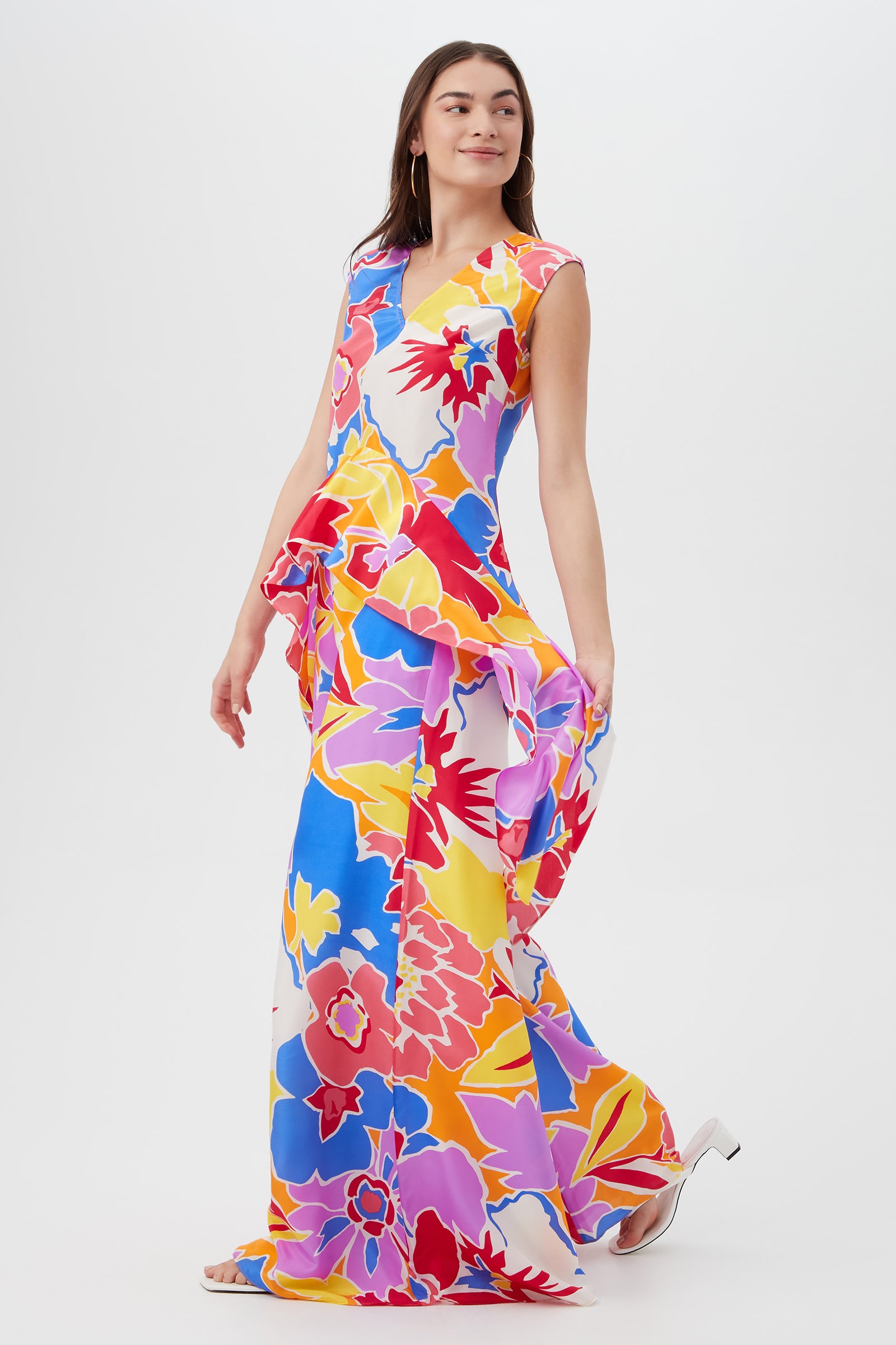 A woman in the Chandelle Top - Soie Paradis, a vibrant sleeveless piece with a bold floral pattern in red, yellow, pink, blue, and white, stands against a plain background holding one side of the top as it flows.