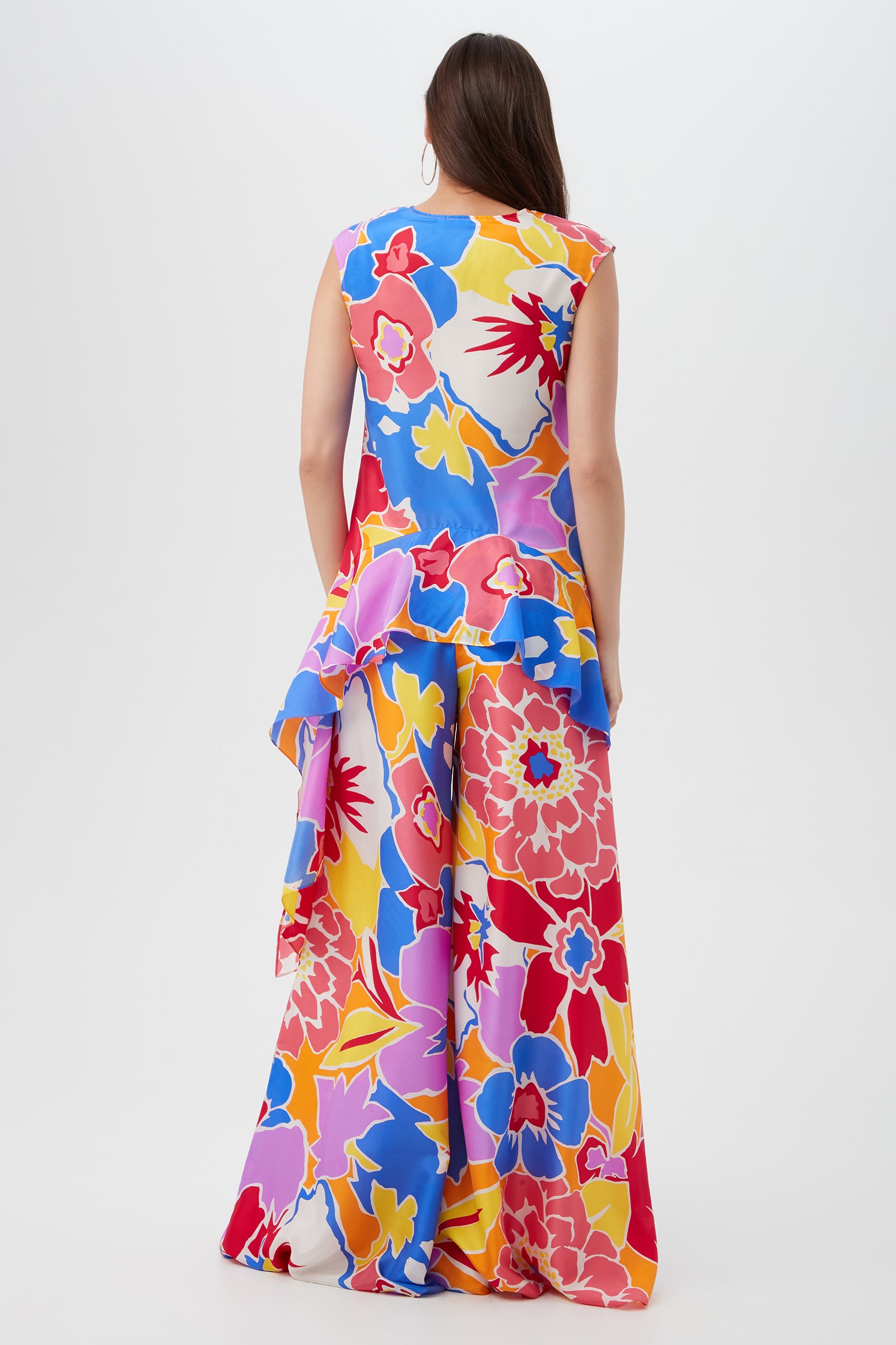 A woman with long brown hair is shown from the back, wearing the Chandelle Top - Soie Paradis, a sleeveless and colorful floral peplum top, set against a plain white background.