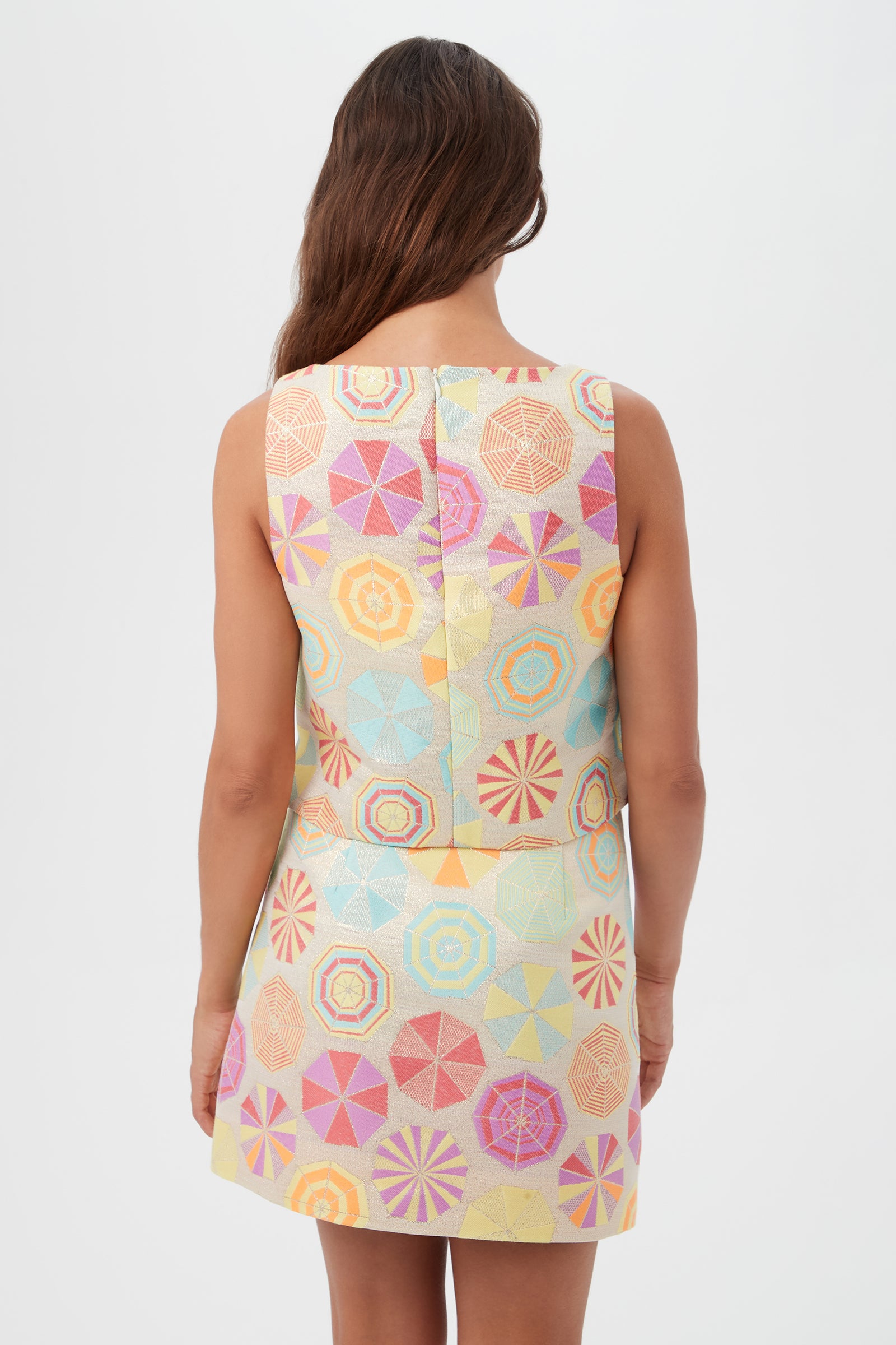 A woman with long brown hair faces away, wearing the sleeveless Cayley 2 Top - Parapluie Jacquard, featuring a pastel geometric umbrella pattern in pink, yellow, blue, and orange. The background is plain white.