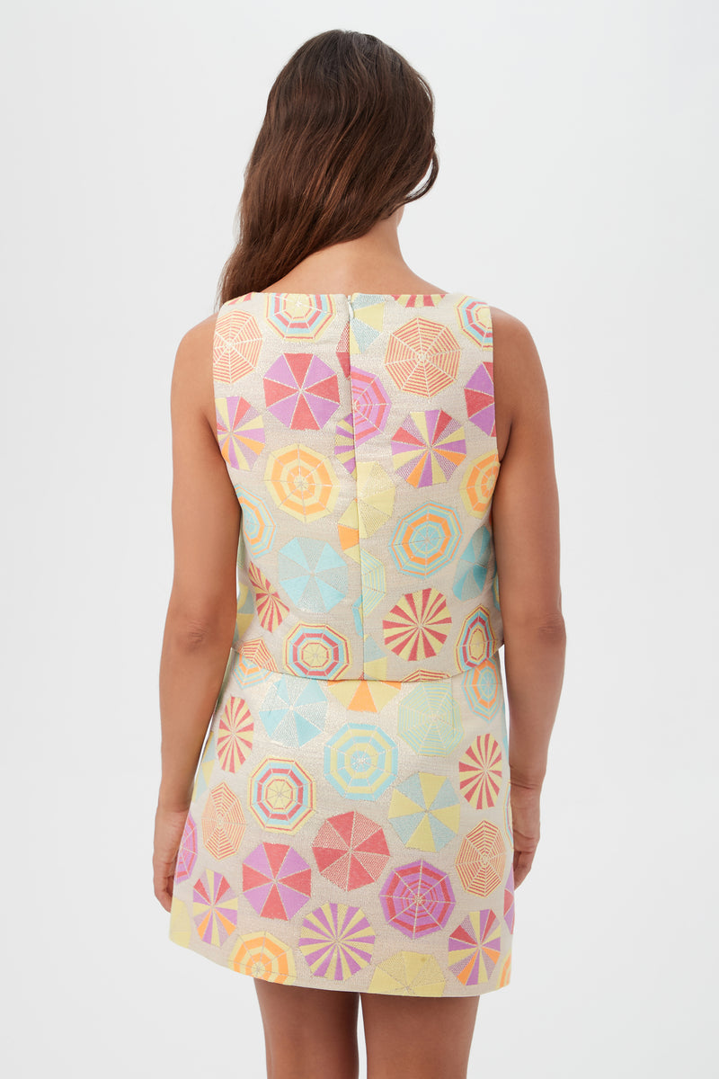A woman with long brown hair faces away, wearing the sleeveless Cayley 2 Top - Parapluie Jacquard, featuring a pastel geometric umbrella pattern in pink, yellow, blue, and orange. The background is plain white.
