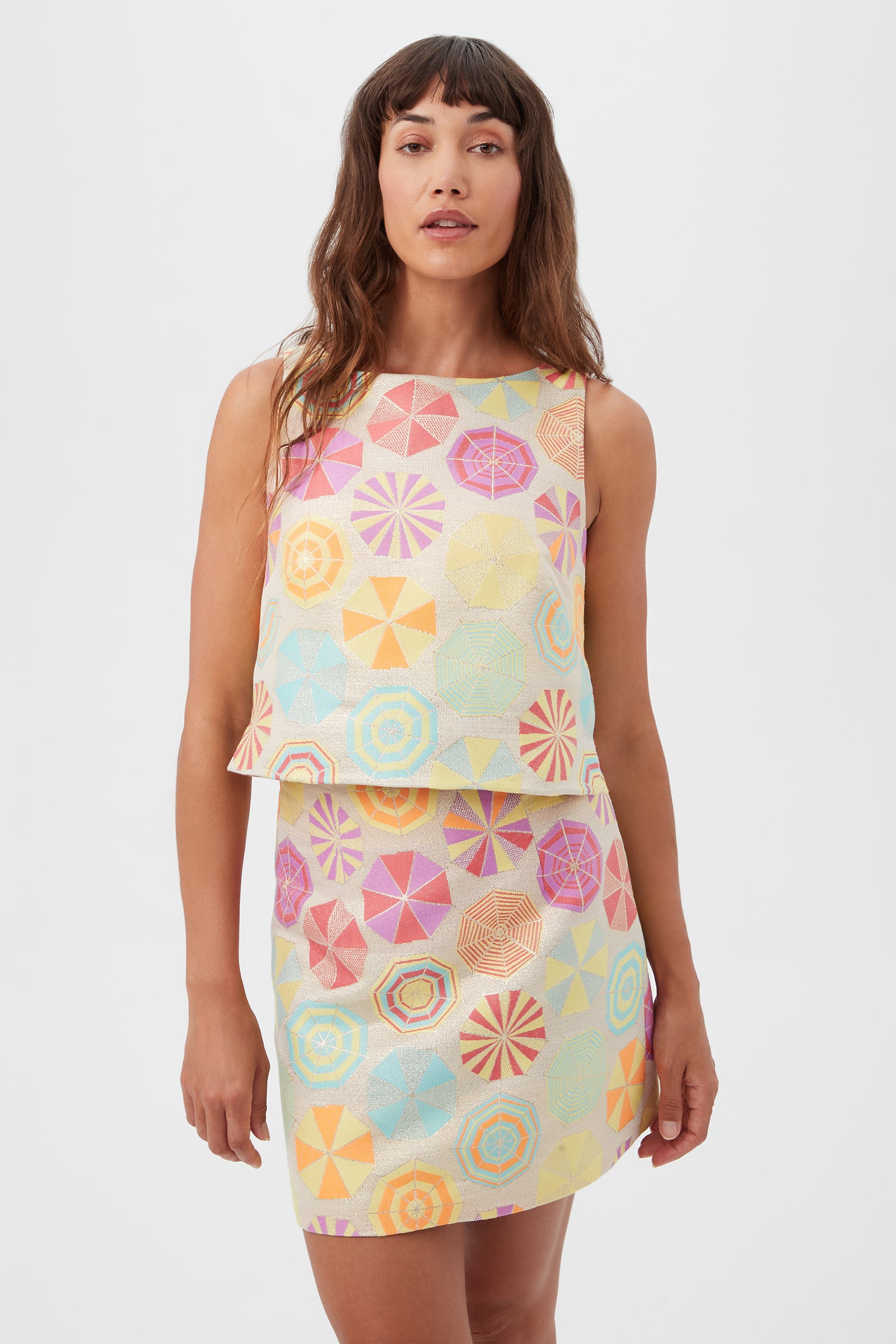 A woman stands against a plain white background wearing the Cayley 2 Top in Parapluie Jacquard, featuring sleeveless styling and colorful geometric patterns.