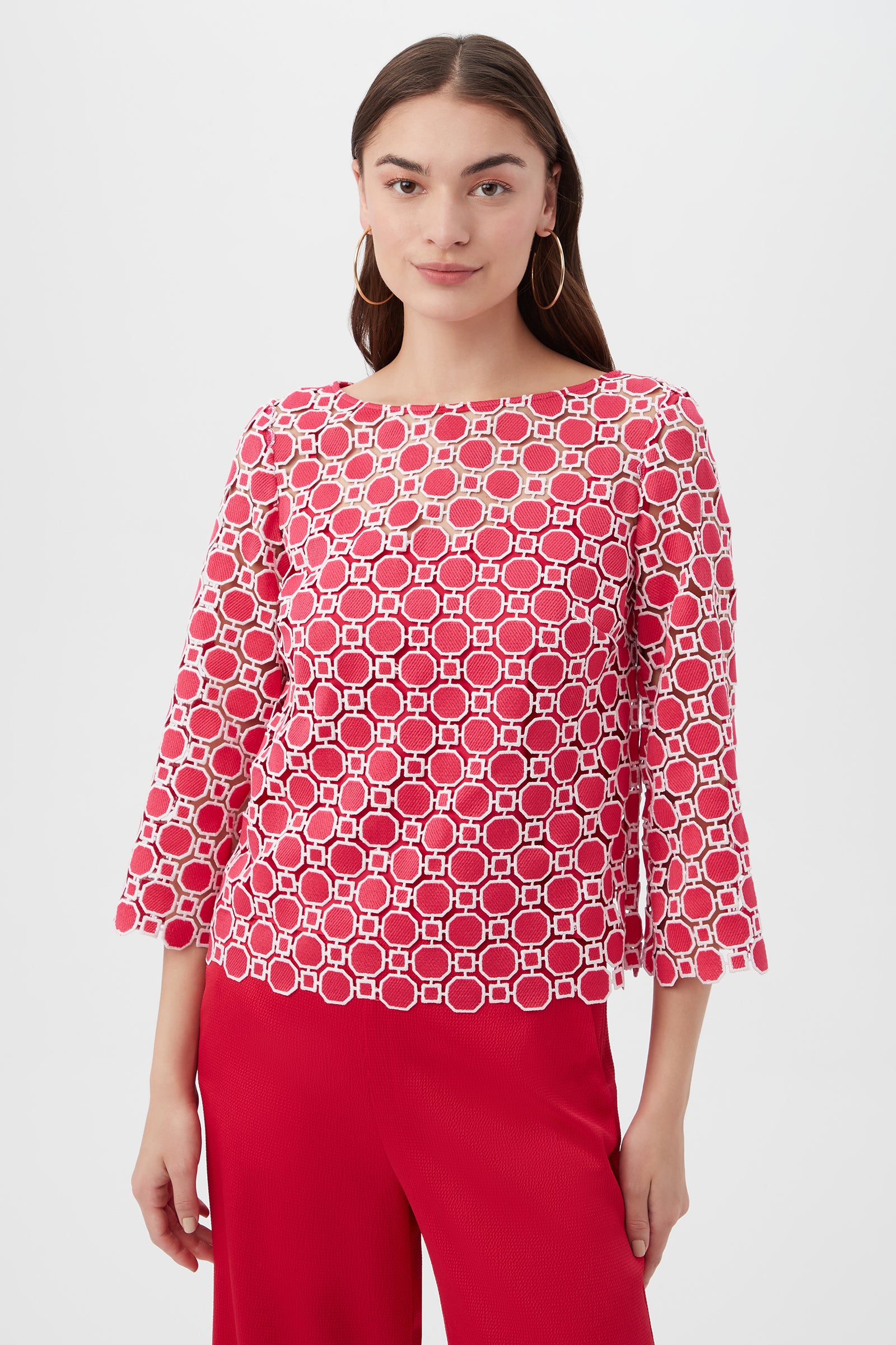 A woman stands against a plain white background wearing the Sun Top in Cannes Coral with 3/4 sleeves and red pants. She has long brown hair, hoop earrings, and a neutral expression.