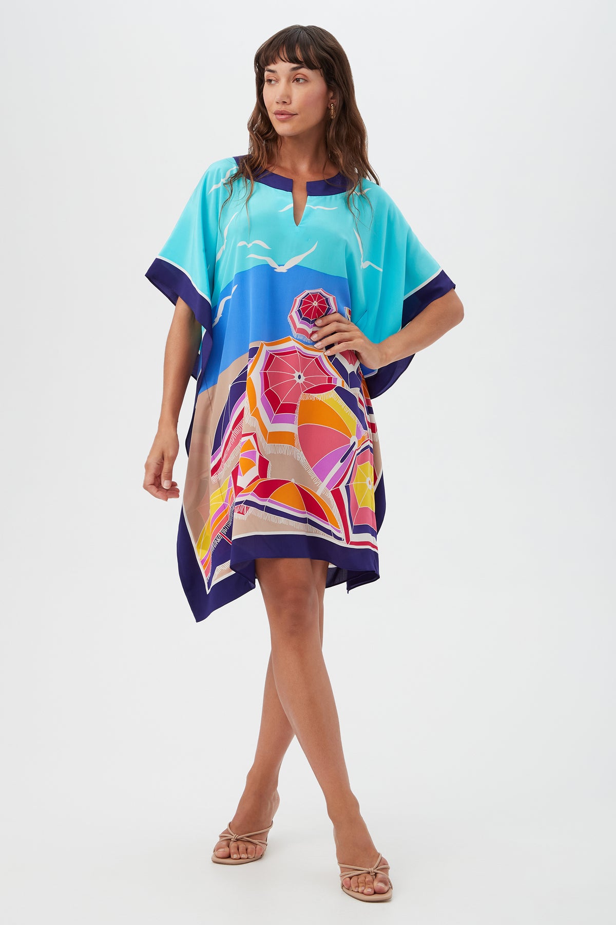 A woman poses barefoot against a white background in Theodora Caftan Dress - Parapluie Silk, a loose multi-colored silk dress with abstract geometric and landscape designs.