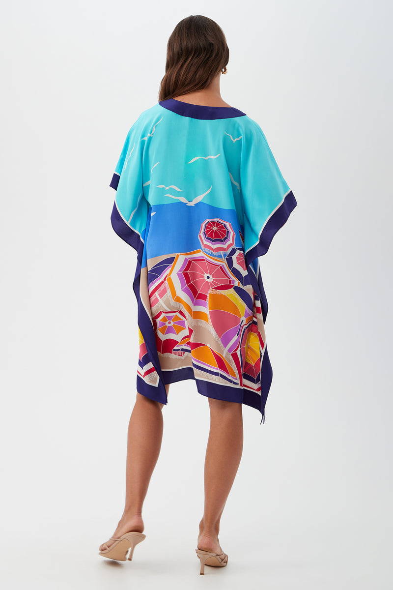 A woman with brown hair faces away, wearing the Theodora Caftan Dress - Parapluie Silk, a loose and colorful silk dress featuring umbrellas, sand, and seagulls. She pairs it with nude heeled sandals against a plain white background.