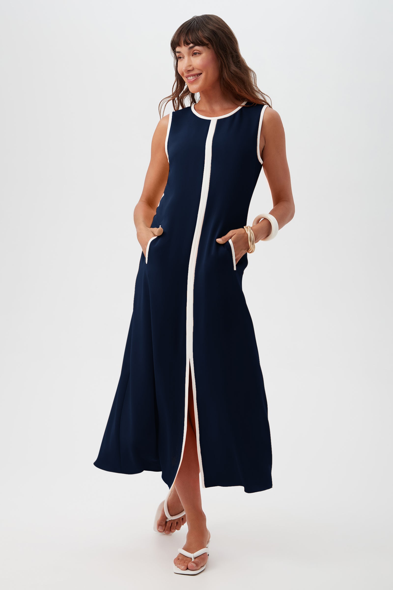 A woman smiles in the Magnifique Dress, a sleeveless navy blue maxi with a central white stripe, white trim, and front pockets. She accessorizes with white sandals and a chunky bracelet, posed against a plain white background.