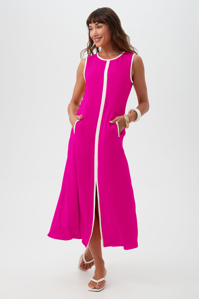 A woman smiles in the Magnifique Dress, a sleeveless bright pink style with white trim along the front and neckline, featuring pockets. She accessorizes with white sandals and a chunky bracelet against a plain white background.