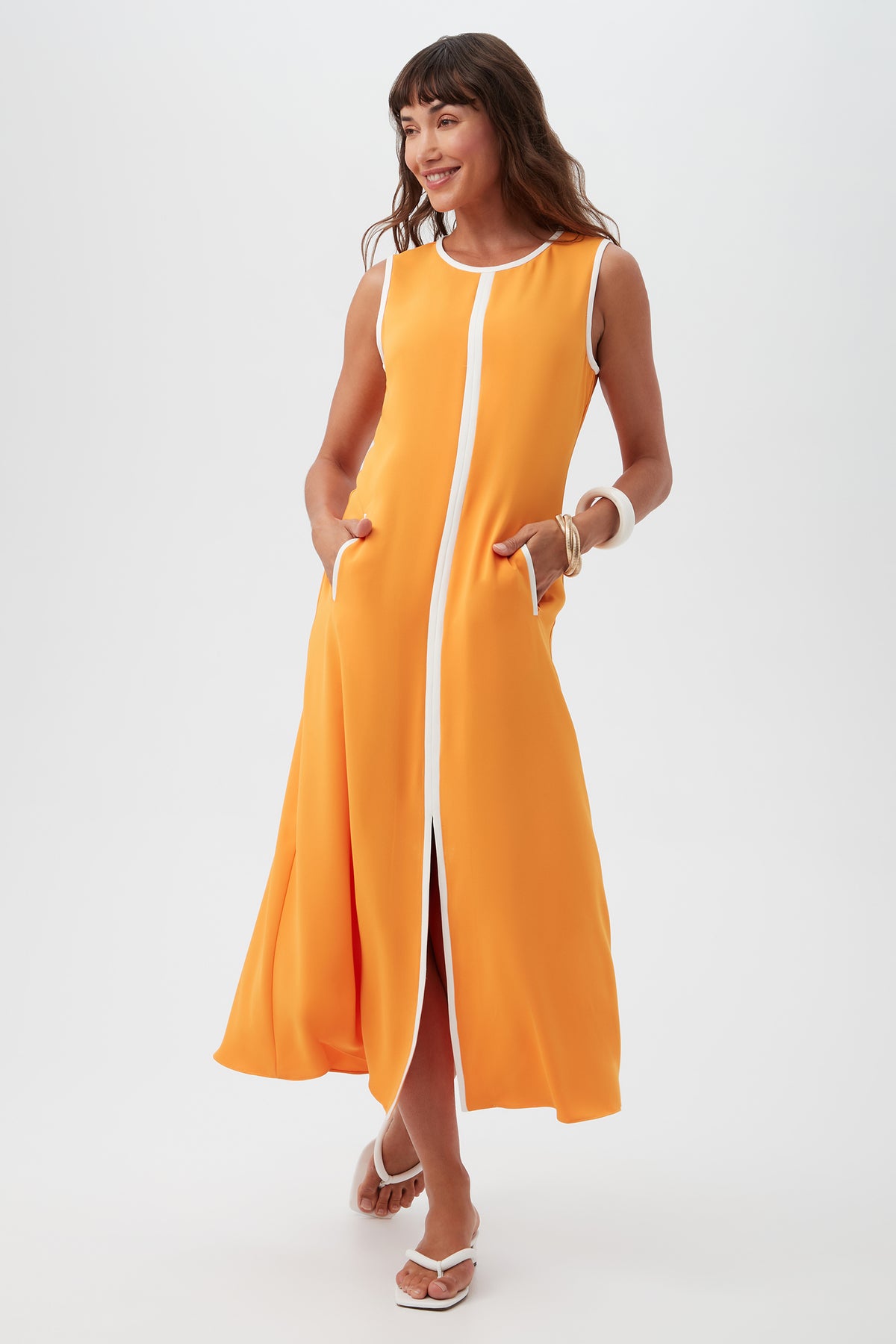A woman models the Magnifique Sleeveless Dress in Cap Clementine, featuring white trim and handy pockets. She stands smiling in sandals against a plain white background.