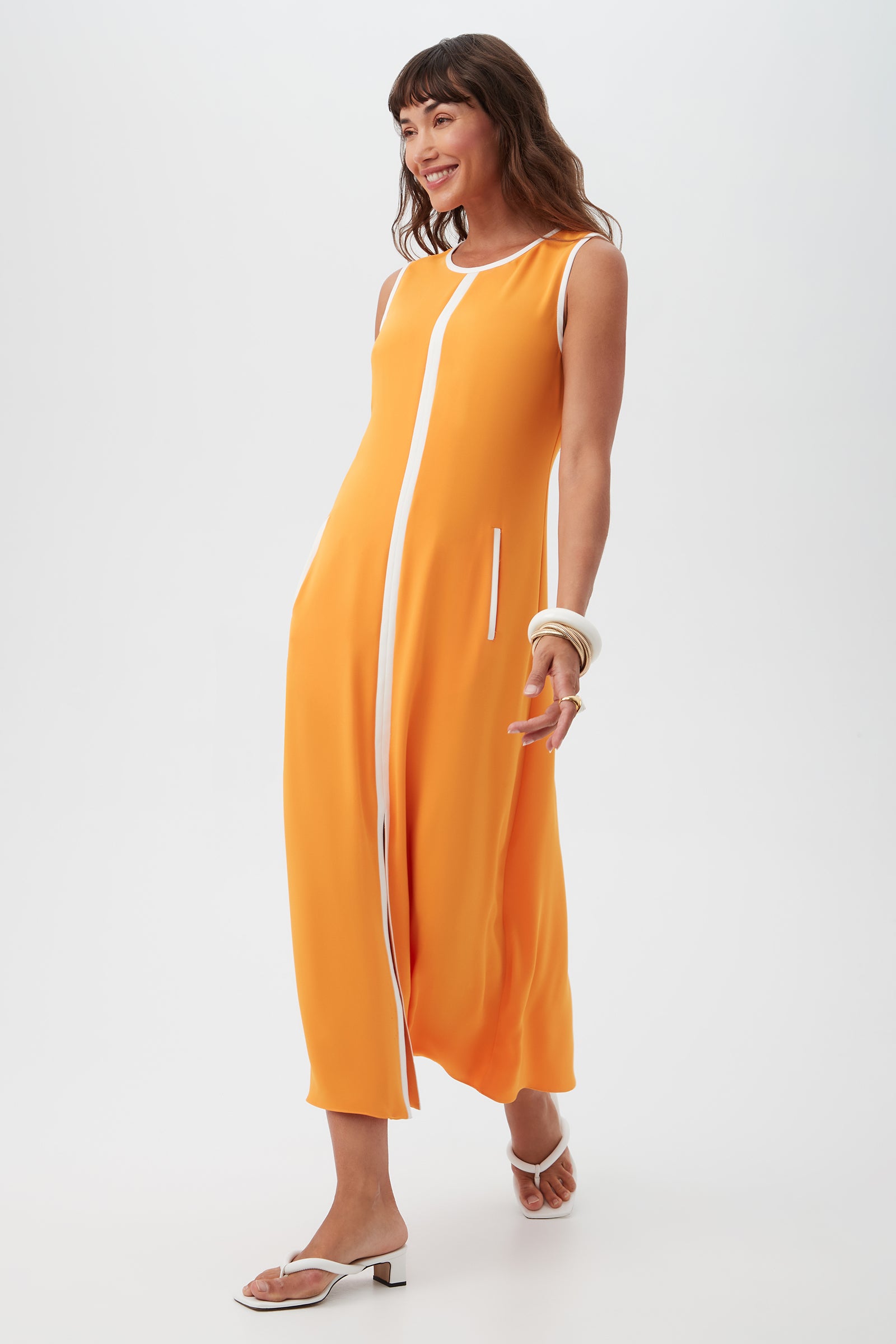 A woman smiles as she walks against a white background, wearing the Magnifique Sleeveless Dress in bright orange with white trim, paired with white sandals and chunky white bracelets.