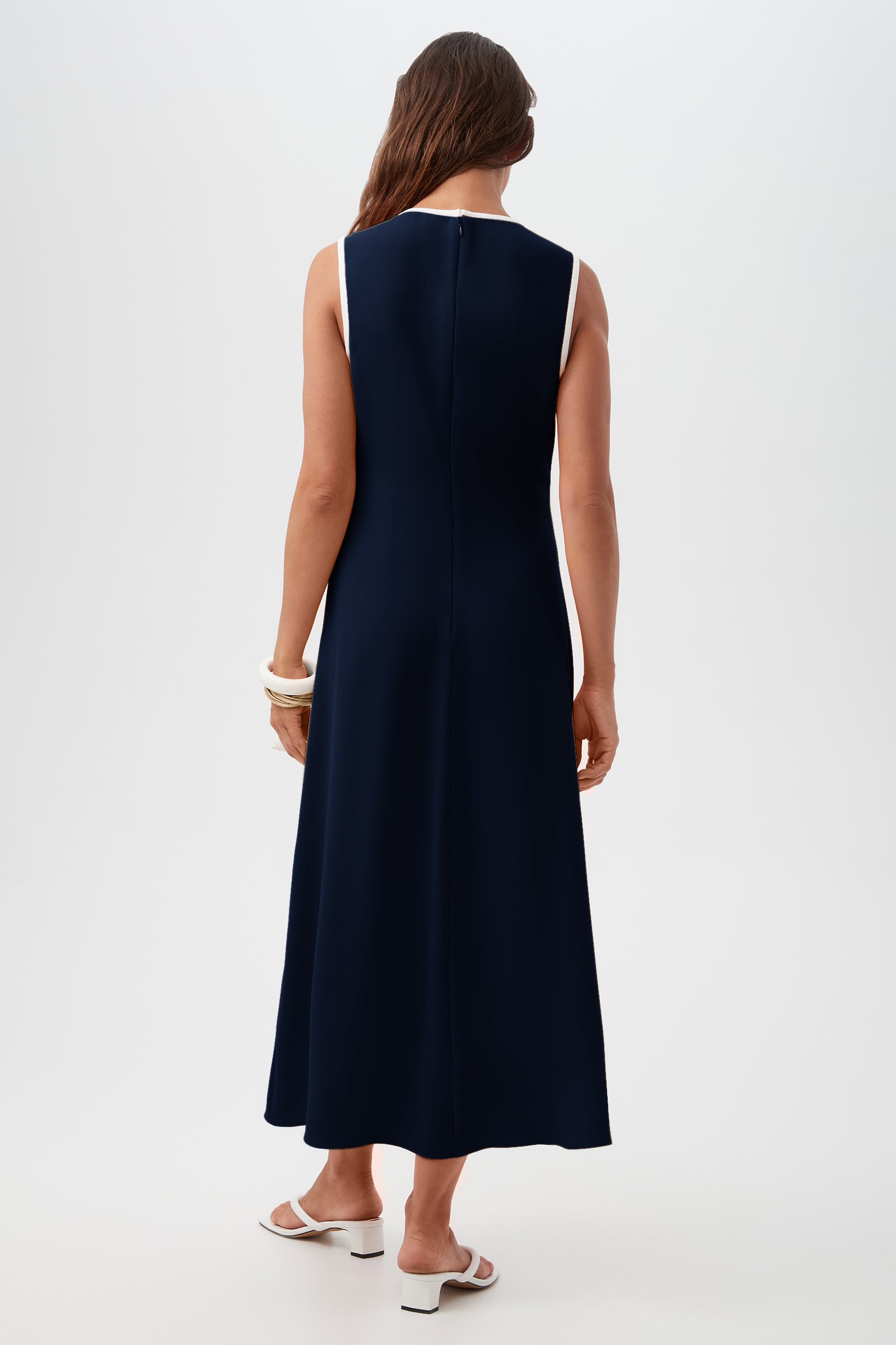 A woman with long brown hair models the Magnifique Dress, a sleeveless, ankle-length navy piece with white trim, paired with white sandals and chunky bracelets. She stands against a plain light background, facing away from the camera.