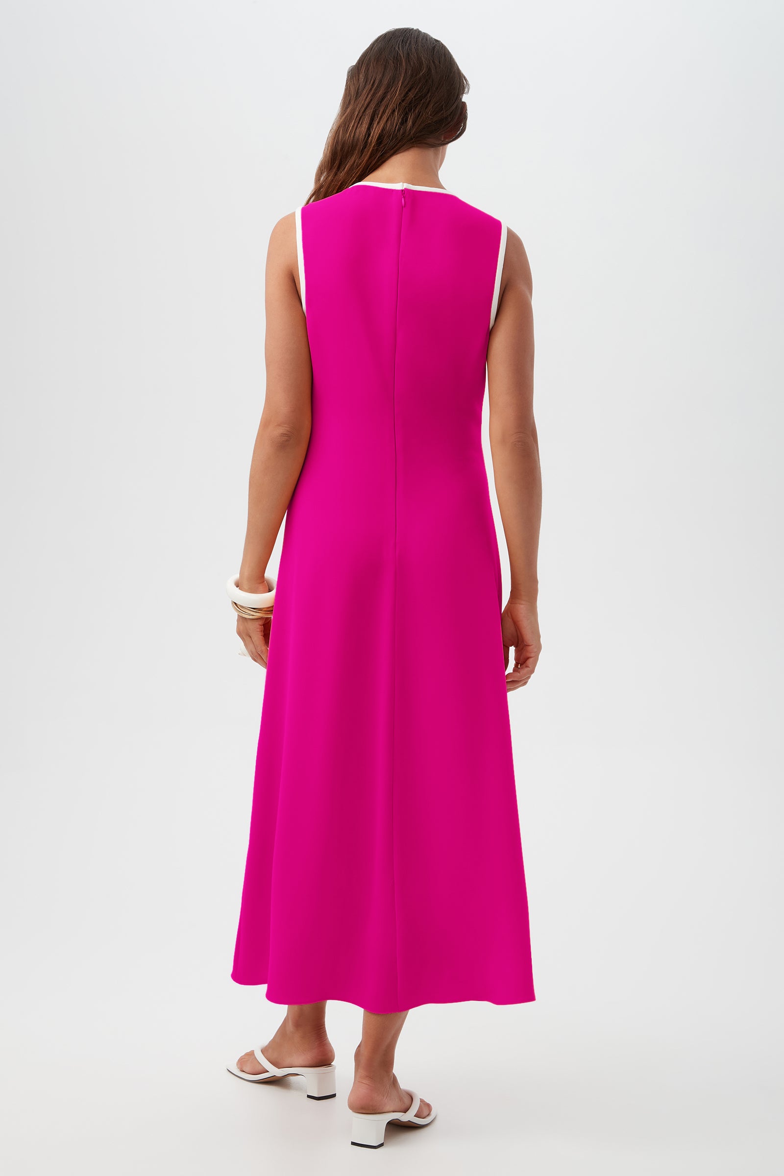 A woman with long brown hair, wearing the Magnifique Dress—a sleeveless, ankle-length bright pink dress with white trim—stands facing away. She pairs it with white sandals and chunky bracelets against a plain white background.