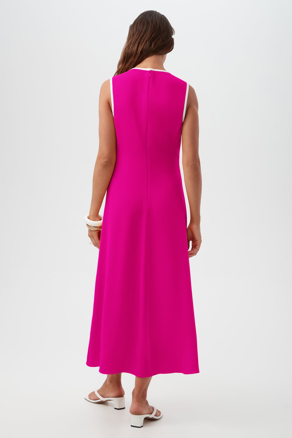 A woman with long brown hair, wearing the Magnifique Dress—a sleeveless, ankle-length bright pink dress with white trim—stands facing away. She pairs it with white sandals and chunky bracelets against a plain white background.