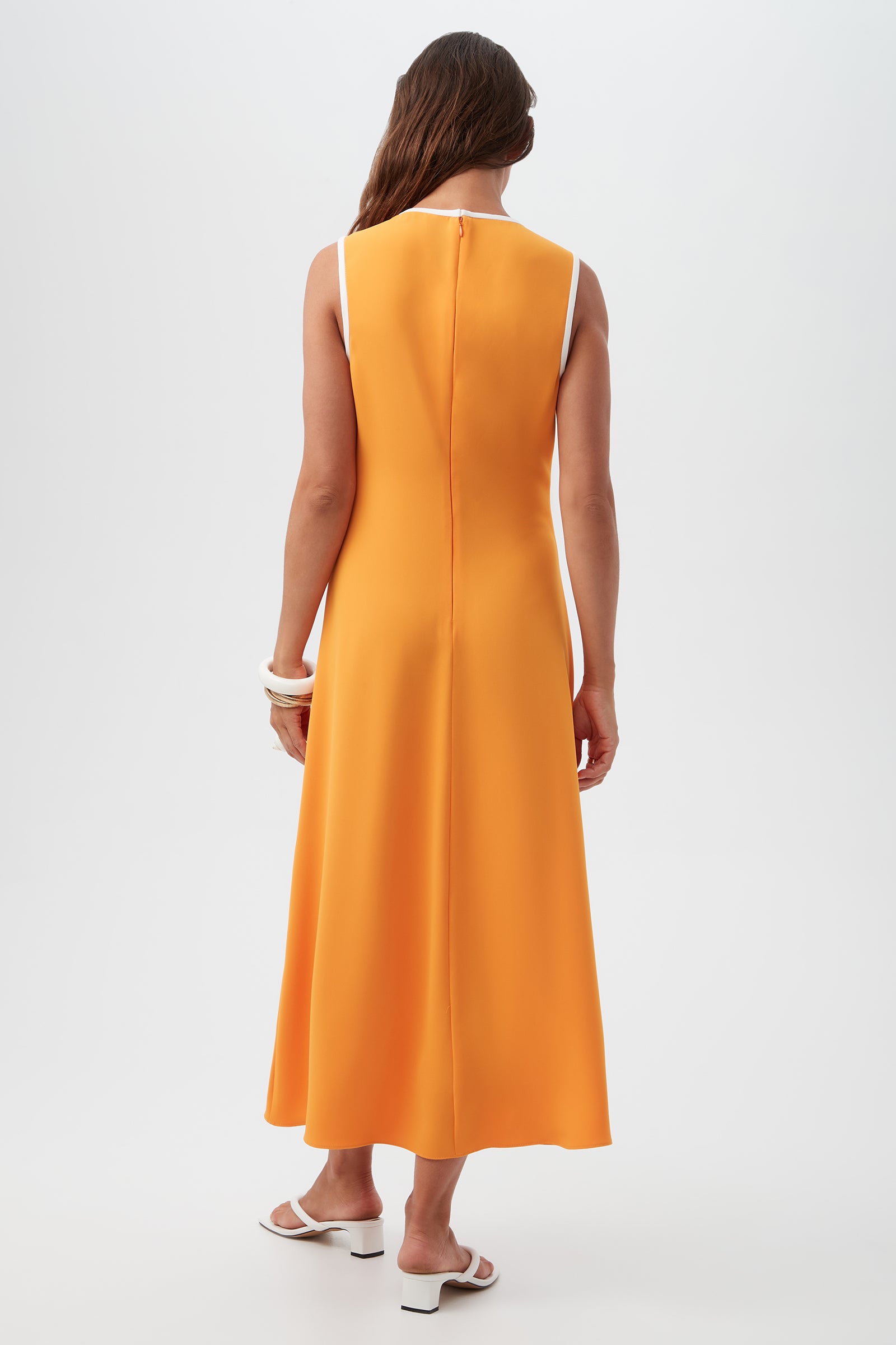A woman with long brown hair models the Magnifique Sleeveless Dress—an ankle-length orange dress with white trim—paired with white sandals and chunky bracelets, standing against a plain white background facing away from the camera.