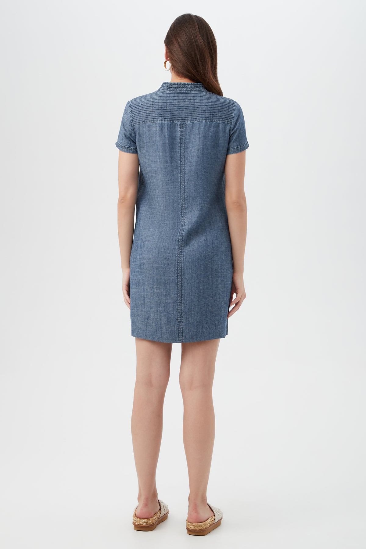 A woman faces away from the camera in a Suntan 2 Short-Sleeve Chambray Dress and brown sandals, with a plain white background.