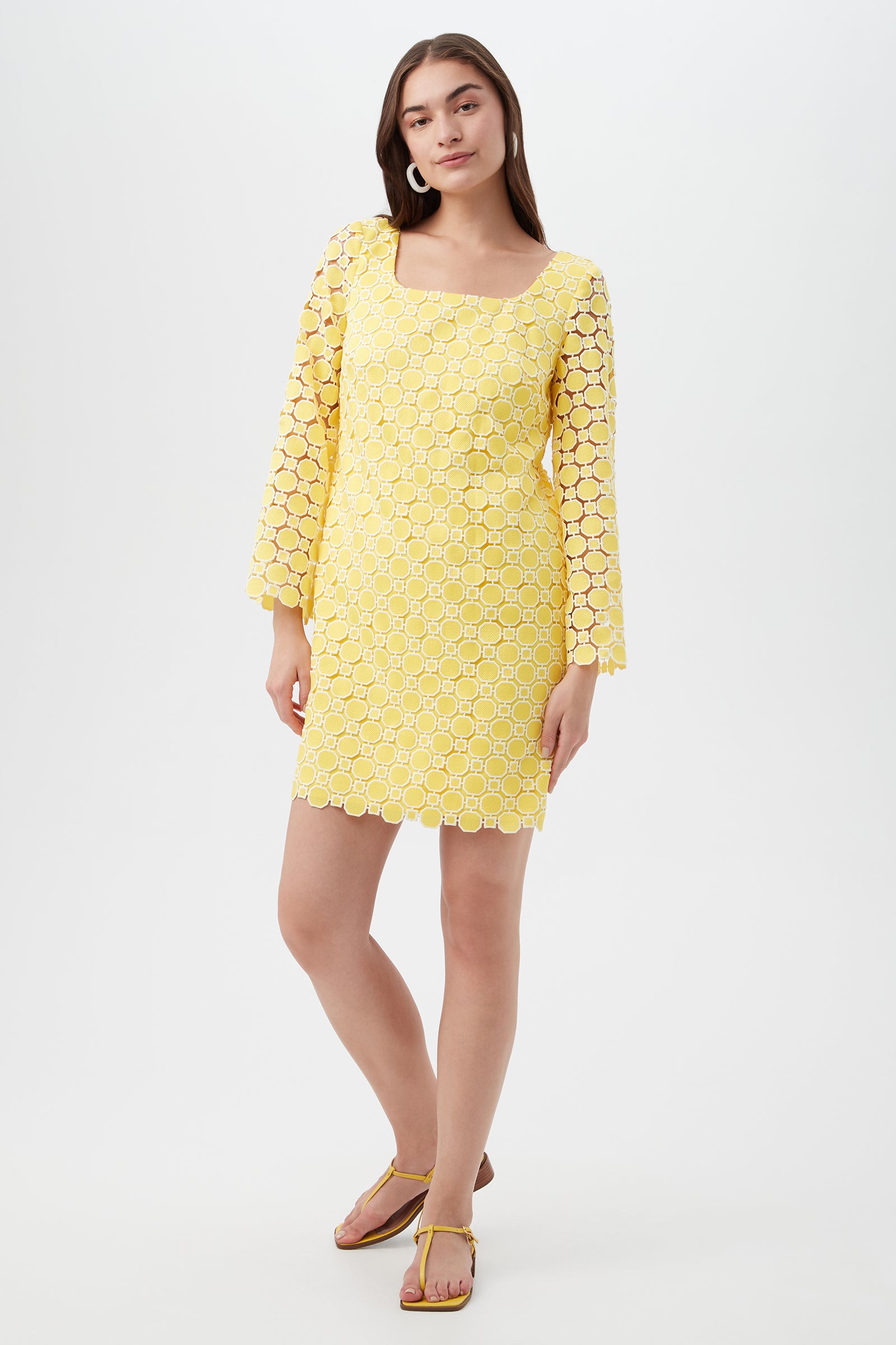 A woman with long brown hair poses with one hand on her hip, wearing the Ariella 2 Long-Sleeve Dress in Lavandou Lemon and matching yellow sandals. The knee-length dress has a square neckline and circular lace pattern.