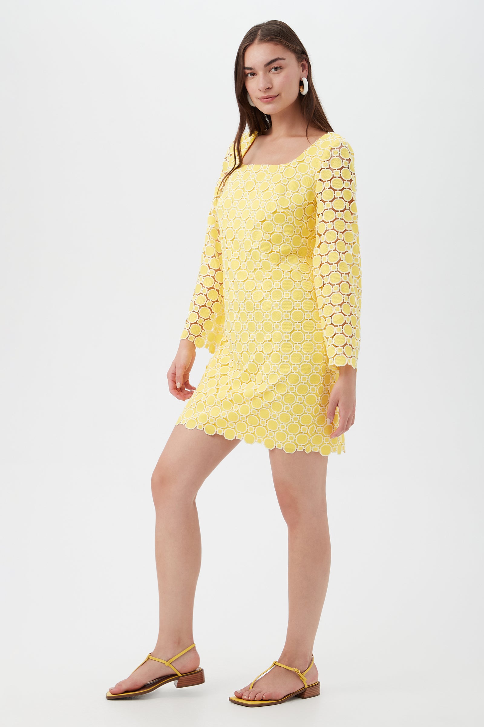 A woman with long brown hair and hoop earrings poses in the Ariella 2 Long-Sleeve Dress—a yellow, crochet lace mini dress with a square neckline—paired with sandals, against a plain white background.