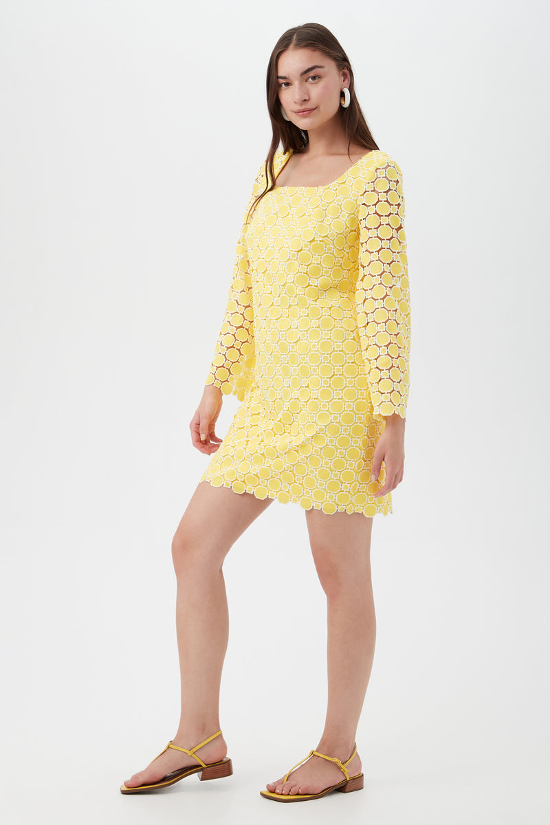 A woman with long brown hair and hoop earrings poses in the Ariella 2 Long-Sleeve Dress—a yellow, crochet lace mini dress with a square neckline—paired with sandals, against a plain white background.