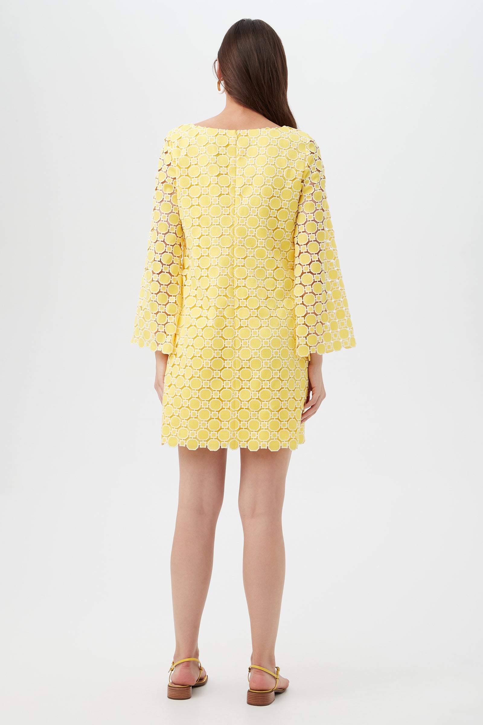 A woman faces away, wearing the Ariella 2 Long-Sleeve Dress—a knee-length yellow dress with circular lace pattern and wide sleeves. She pairs it with sandals, her straight brown hair down, standing against a plain light background.