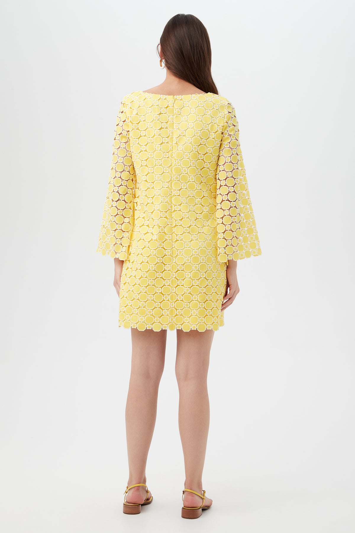 A woman faces away, wearing the Ariella 2 Long-Sleeve Dress—a knee-length yellow dress with circular lace pattern and wide sleeves. She pairs it with sandals, her straight brown hair down, standing against a plain light background.